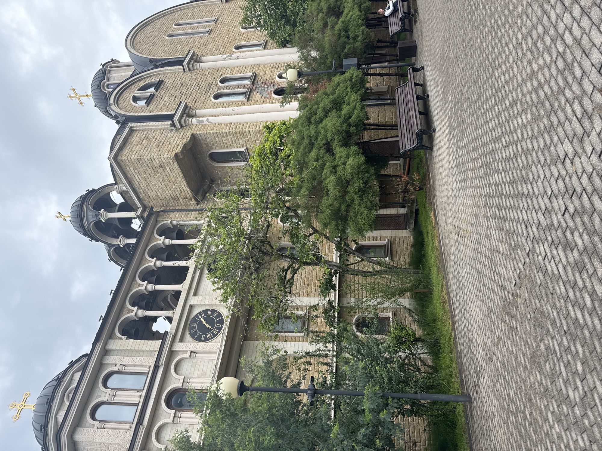The image shows a building in the city of Sochi, Russia. The building is likely a church, as evidenced by the presence of crosses on the domes. The facade of the building is constructed of light-colored stone. A clock is visible on one side of the building. Benches and trees are located adjacent to the building, and a paved walkway leads along the side. The sky appears overcast.