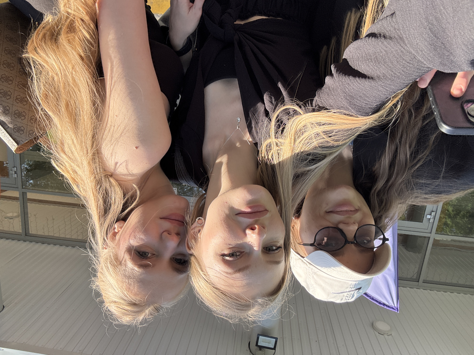 Three young women are posed close together, likely taking a selfie. The woman on the left has long, blonde hair, pale skin, and is wearing a dark top. The woman in the center has shoulder-length blonde hair, pale skin, a silver necklace, and is wearing a black top tied at the front. The woman on the right has long, light brown hair with blonde highlights, is wearing sunglasses, a white baseball cap, and a gray textured top. The background shows a white-painted building with windows, indicating an exterior setting. A purple banner or flag is visible behind the woman on the right. The location is Perm, Russia.