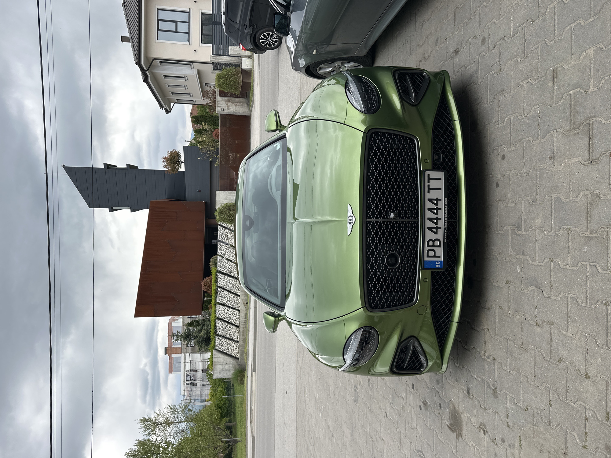 The image shows a green Bentley automobile parked on a paved surface. The car's license plate reads "PB 4444 TT," indicating registration in Bulgaria. Adjacent to the Bentley is another dark-colored car. Behind the cars is a modern architectural structure with rust-colored metal cladding. Further in the background are buildings and trees under a cloudy sky. The location appears to be an urban area in Plovdiv, Bulgaria.