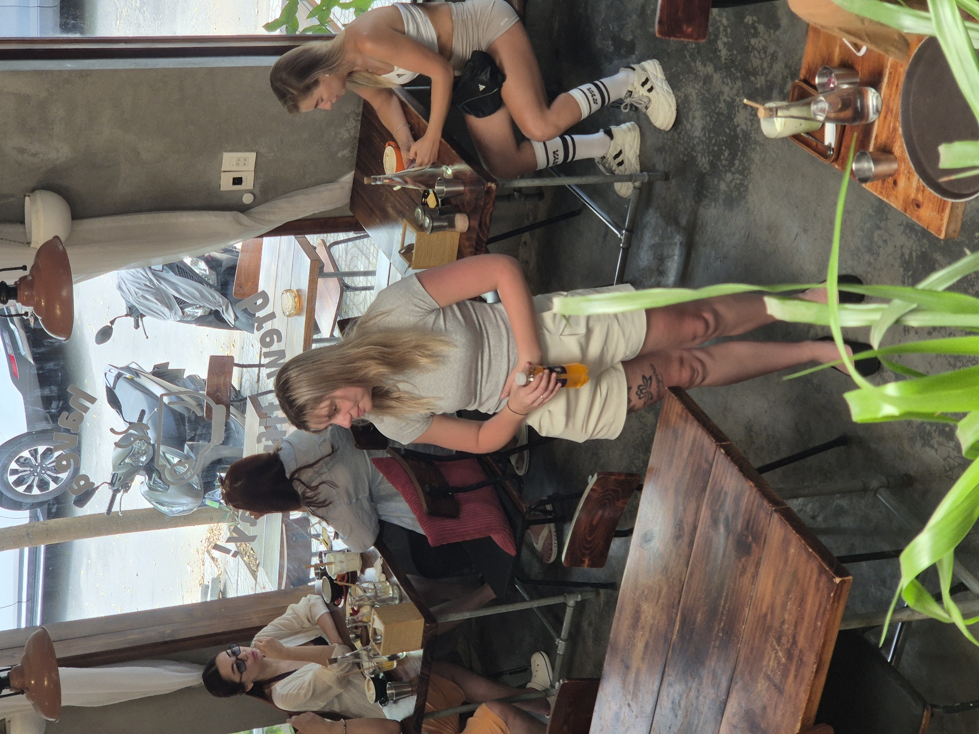 The image shows a group of people seated at tables inside a cafe. In the foreground, a woman with blonde hair and wearing a grey t-shirt and beige shorts stands holding a bottle of orange liquid. She has a tattoo on her left thigh. To her right, another woman with blonde hair wearing a white sports bra and grey shorts is seated at a table, with her legs crossed. She is wearing white socks with black stripes and white sneakers.
In the background, a woman with dark hair and sunglasses is seated at a table, wearing a white cardigan over a dark top. To her right, a woman with dark hair is wearing a light blue shirt. The cafe has a rustic interior with wooden tables and chairs. Through the large windows, a street scene is visible, including a motorcycle and a parked car. The environment appears to be a street-level establishment in a city. The presence of a motorcycle and the general atmosphere suggests an urban setting in Southeast Asia. While not explicitly stated, the visual cues are consistent with a cafe in Đà Nẵng, Vietnam.