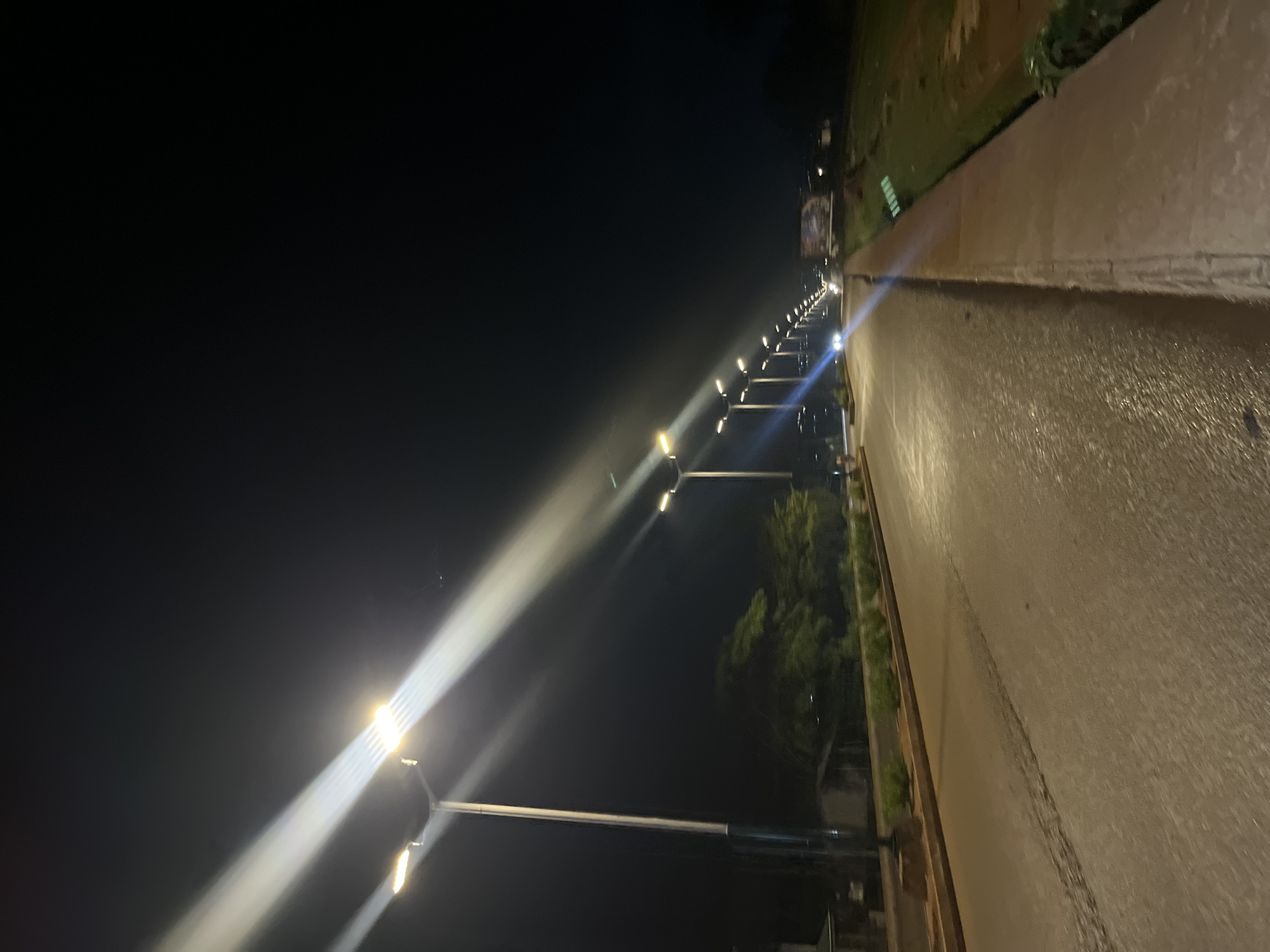 The image depicts a nighttime scene on a road in Bukuru, Nigeria, likely a street lined with modern lampposts. The road is paved and appears wet, reflecting the bright lights from the lampposts that illuminate the scene with strong beams. The sky is dark, indicating nighttime. On the left side of the road, there are some buildings and trees visible. On the right side, there is a raised embankment or sidewalk covered in grass and what appears to be a ditch or drainage channel. Further down the road, a few vehicles can be seen, their headlights cutting through the darkness. The overall impression is one of a well-lit urban or suburban street at night.