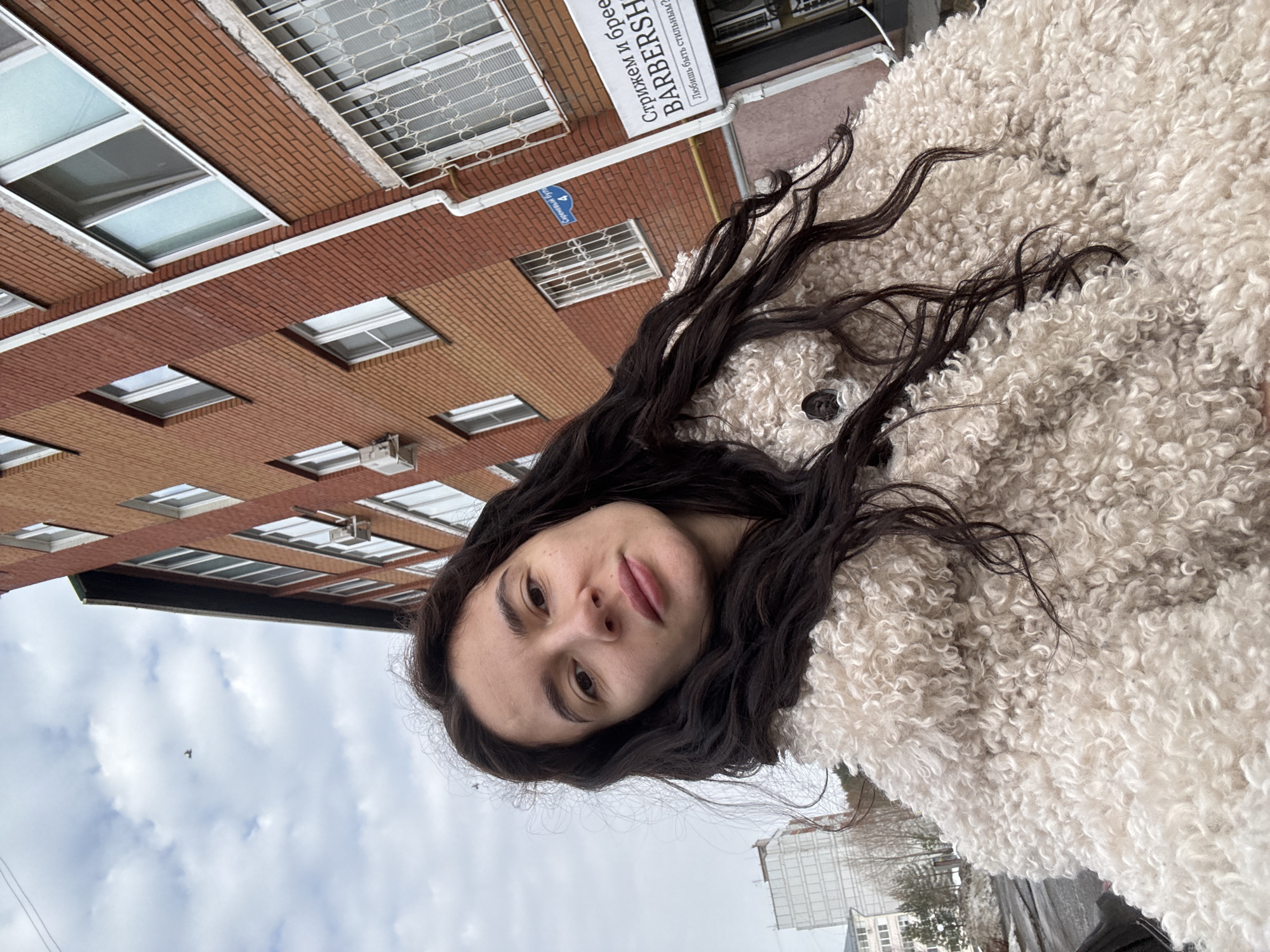 A female subject is depicted in a close-up selfie, looking directly into the camera with a neutral expression. She has long, dark, wavy hair and is wearing a light-colored, cream or off-white, textured faux fur coat. The scene is located at Syrenevyy Bulvar (Сиреневый бул.) 4 in Chaikovsky, Perm Krai, Russia.
The background features a multi-story brick building, predominantly red and orange, with numerous windows. Some windows on the lower levels are protected by decorative metal security grilles. A white downspout runs vertically along the building's facade. An external air conditioning unit is mounted on the brick wall. A commercial sign on the building indicates a "BARBERSHOP" with the Russian text "Стрижем и бреем" (We cut and shave) and "Любишь быть стильным?" (Love to be stylish?).
To the left of the subject, a more distant, lighter-colored tall building is visible, contrasting with the brick structure. The sky is overcast with visible clouds, and a single bird is captured in flight. The ground partially visible in the lower left suggests a paved surface with dark, possibly wet or melted snow patches. Sparse, bare vegetation is visible in the distant background.