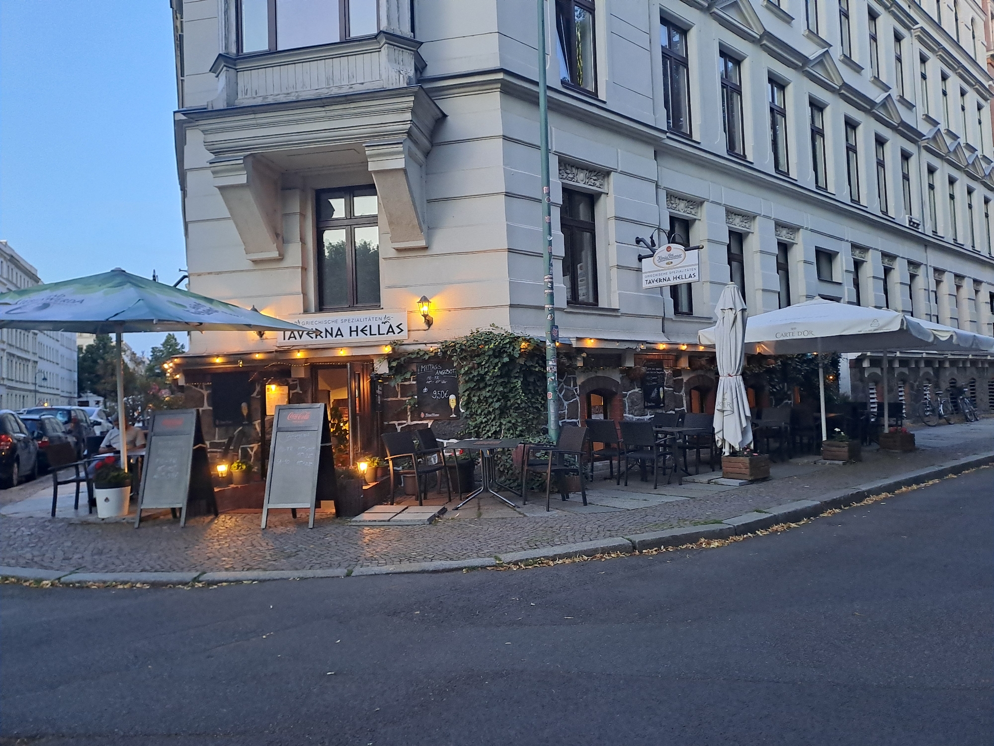 The image shows the exterior of a corner building with a restaurant named "Taverna Hellas" on the ground floor, located in Leipzig, Germany. The building features ornate architectural details, including a bay window on the upper floor and decorative molding. The restaurant has outdoor seating with umbrellas, tables, and chairs set up on the cobblestone sidewalk. Two A-frame signs stand in front of the restaurant, one advertising Coca-Cola. Cars are parked along the street in the background. The scene appears to be set in the late afternoon or early evening, with warm lighting from the restaurant and streetlights.