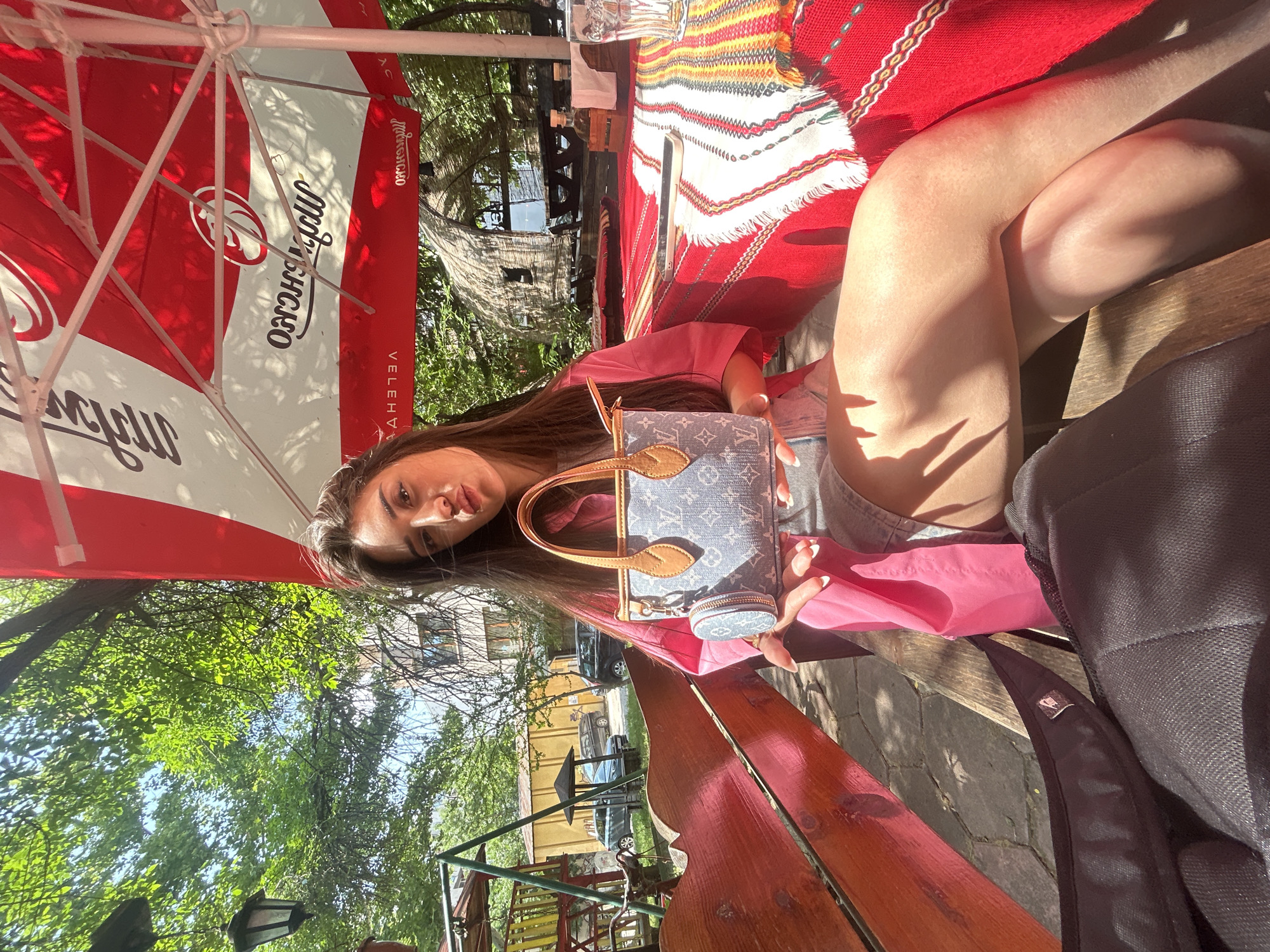 The image, oriented vertically (rotated 90 degrees clockwise from standard portrait), depicts a young woman relaxing outdoors at a cafe or restaurant setting, likely in Plovdiv, Bulgaria, on a bright, sunny day. She has long, dark hair and is wearing a pink top and denim shorts. She looks directly towards the camera with a neutral expression, holding a light blue Louis Vuitton monogram handbag with brown leather trim; a matching small coin purse is attached to the bag.
She is seated on a reddish-brown wooden bench at a matching wooden table, which is covered with a vibrant red textile featuring traditional-looking striped patterns in yellow, white, and other colors. A black backpack is partially visible next to her on the bench. Above her, a large red and white outdoor umbrella provides shade. The umbrella displays mirrored and upside-down text, clearly showing "Милѣчскоро" and "ВЕЛЕХВА" (Milechskoro and Velehva).
The scene is well-lit by natural sunlight, casting distinct shadows on her legs and the paved ground, indicative of clear weather. In the background, lush green trees and foliage suggest a natural, possibly garden-like environment. Beyond the greenery, parts of buildings, including an older light-colored structure, and parked cars are visible. The overall atmosphere is relaxed and summery.
