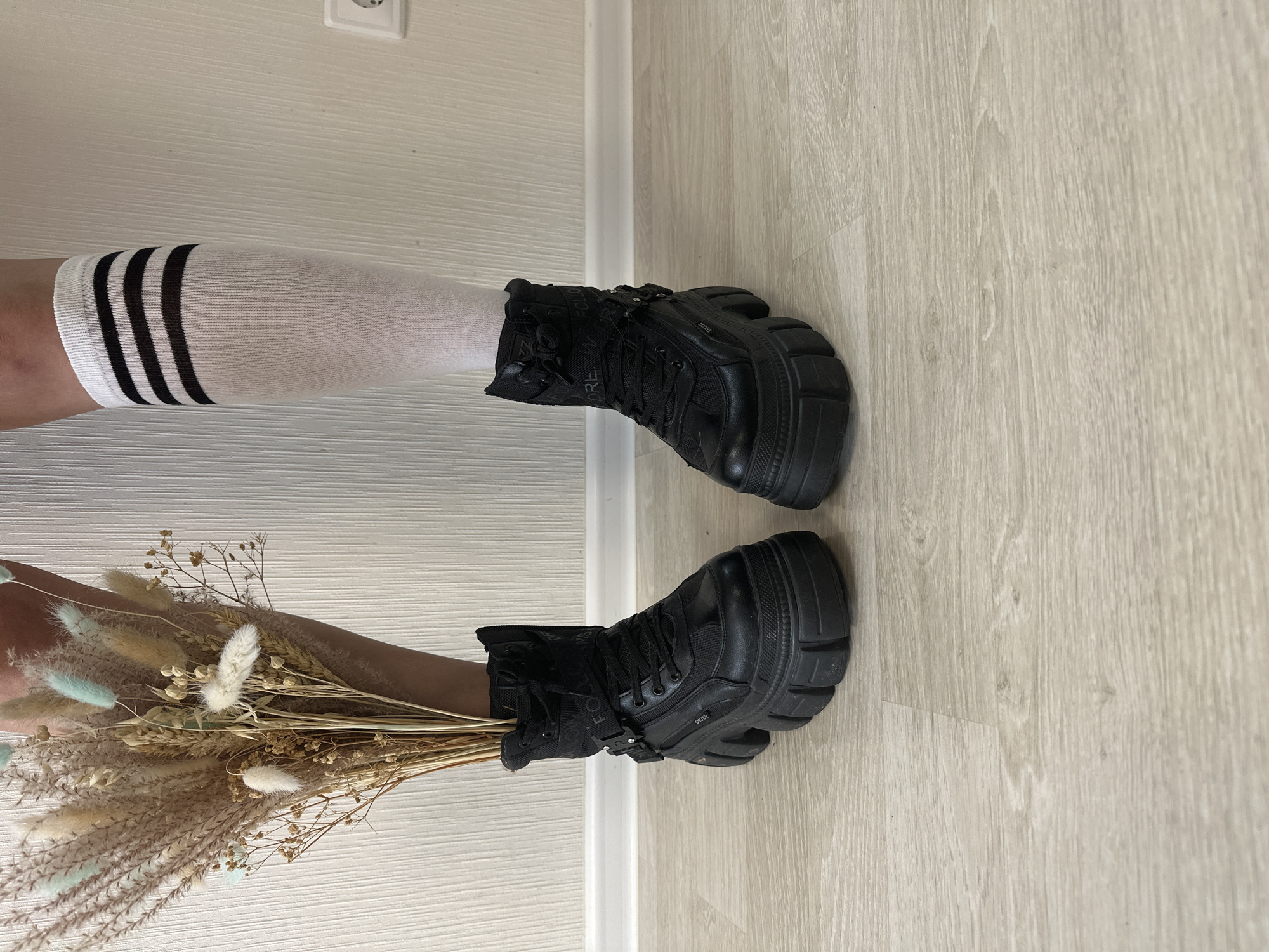 The image shows a close-up of two legs wearing black platform boots. The leg on the left is adorned with a white sock featuring three black stripes, while the leg on the right holds a bouquet of dried flowers. The background consists of a light-colored wall and floor. There is no information present in the image to indicate the city of Izhevsk, Russia.