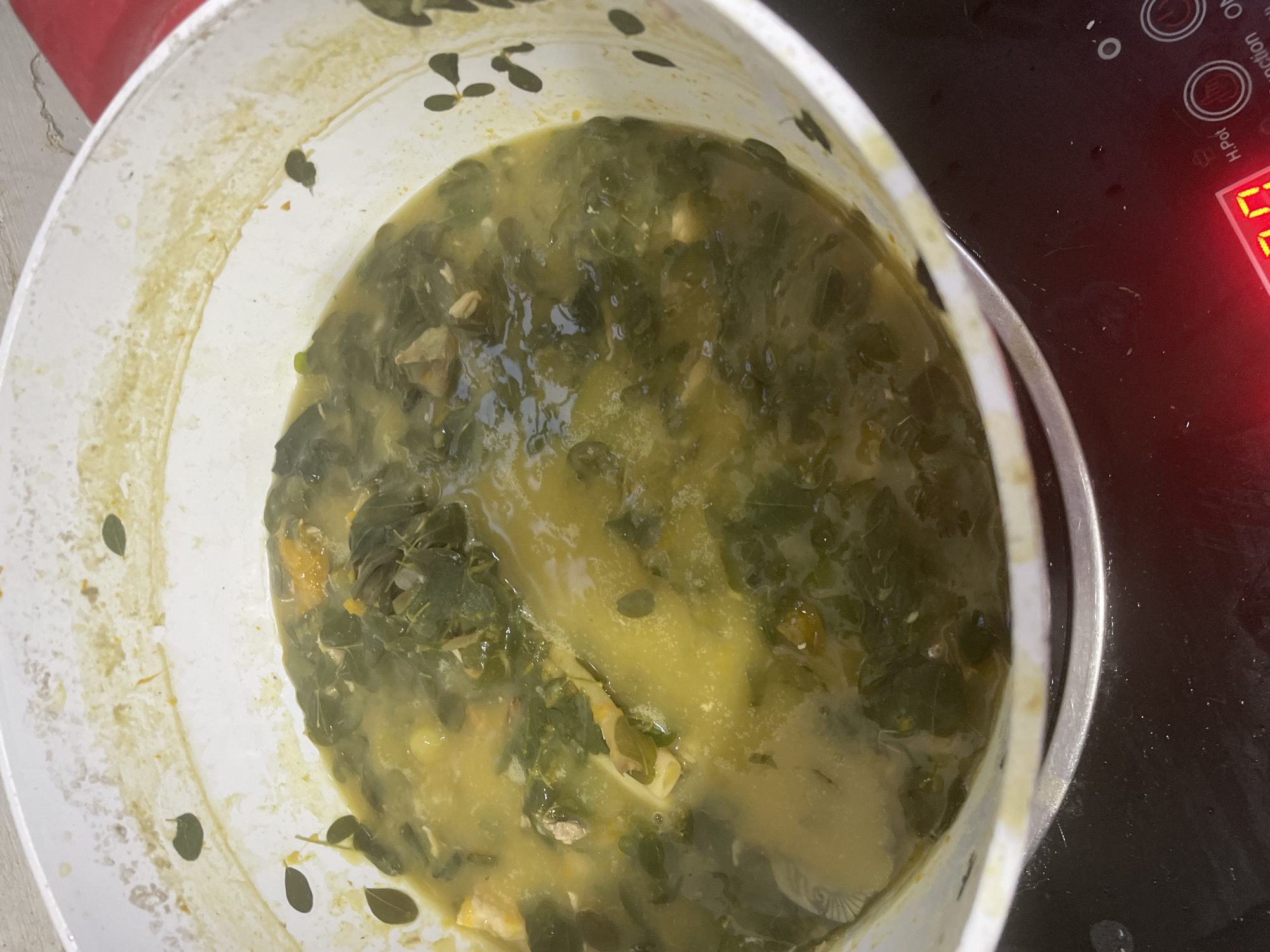 An overhead shot captures a large white pot filled with a green leafy stew or soup, positioned on a black cooktop. The pot contains a thick, yellowish-green broth, generously mixed with numerous small, dark green leaves, strongly resembling malunggay (moringa). Visible within the stew are chunks of light brown meat and softer, yellowish pieces, possibly squash.
The white rim of the pot and its upper exterior show splashes and residue from cooking. To the right of the pot, part of a black induction cooker is visible. It features circular control buttons labeled "ON," "Function," and "H.Pot." A red digital display on the cooktop shows partial numbers, appearing as "0" or "U," indicating it is active.
The setting is an indoor kitchen, with the food being either cooked or kept warm on the stovetop. No individuals are present in the image. The dish appears hearty and fresh, consistent with traditional Filipino cuisine, particularly in the context of Davao City, Philippines.