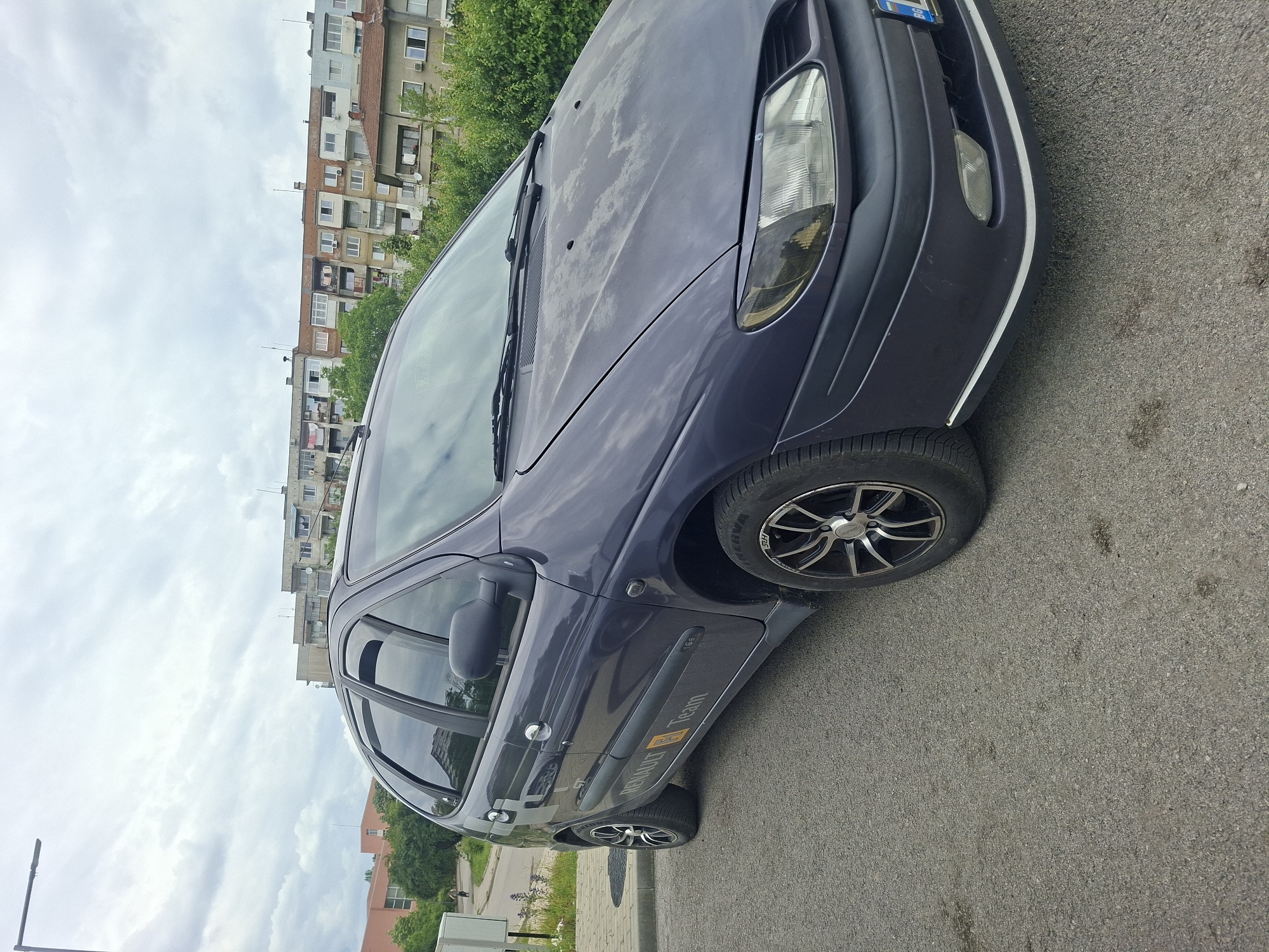 A medium-high angle shot of a dark blue Renault Scenic team car parked on an asphalt street, facing right. The front end of the car is in the upper right of the frame. Its right-side front and rear tires are in view. The car is parked parallel to a sidewalk and the curb. Behind the car is a building. The sky above is mostly cloudy.