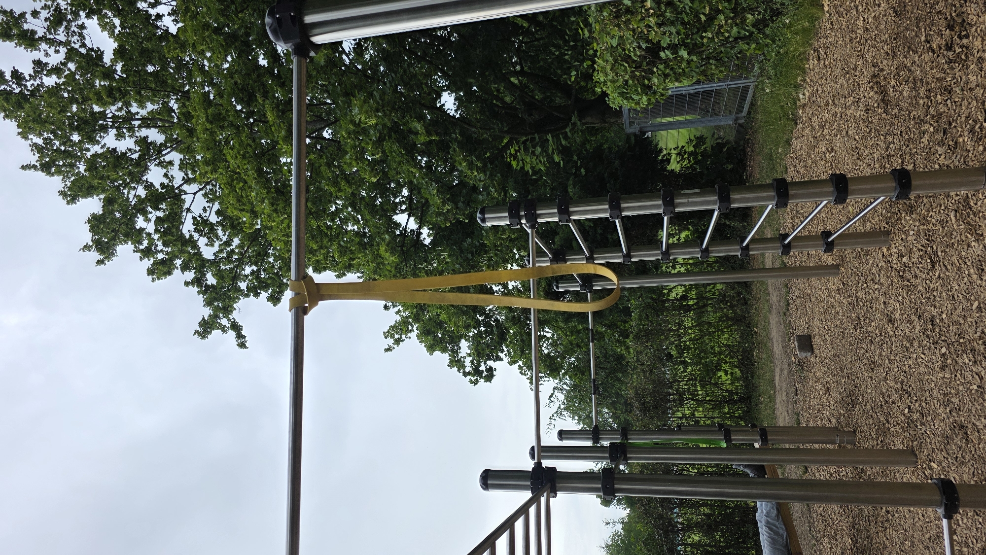 An outdoor exercise area features a bright yellow resistance band hanging from a horizontal metal bar. The band is looped and dangles downwards, creating a U-shape. The bar is part of a larger structure made of shiny metal poles and bars, including a ladder-like section on the right. The ground is covered in light brown wood chips. In the background, a dense green tree with full foliage dominates the upper right of the frame, with a cloudy sky above. To the left of the exercise equipment, a line of green bushes and trees is visible, and further to the left, a portion of what appears to be a bench or table covered with a gray sheet can be seen. A dark metal fence is visible in the far background on the right, partially obscured by foliage.