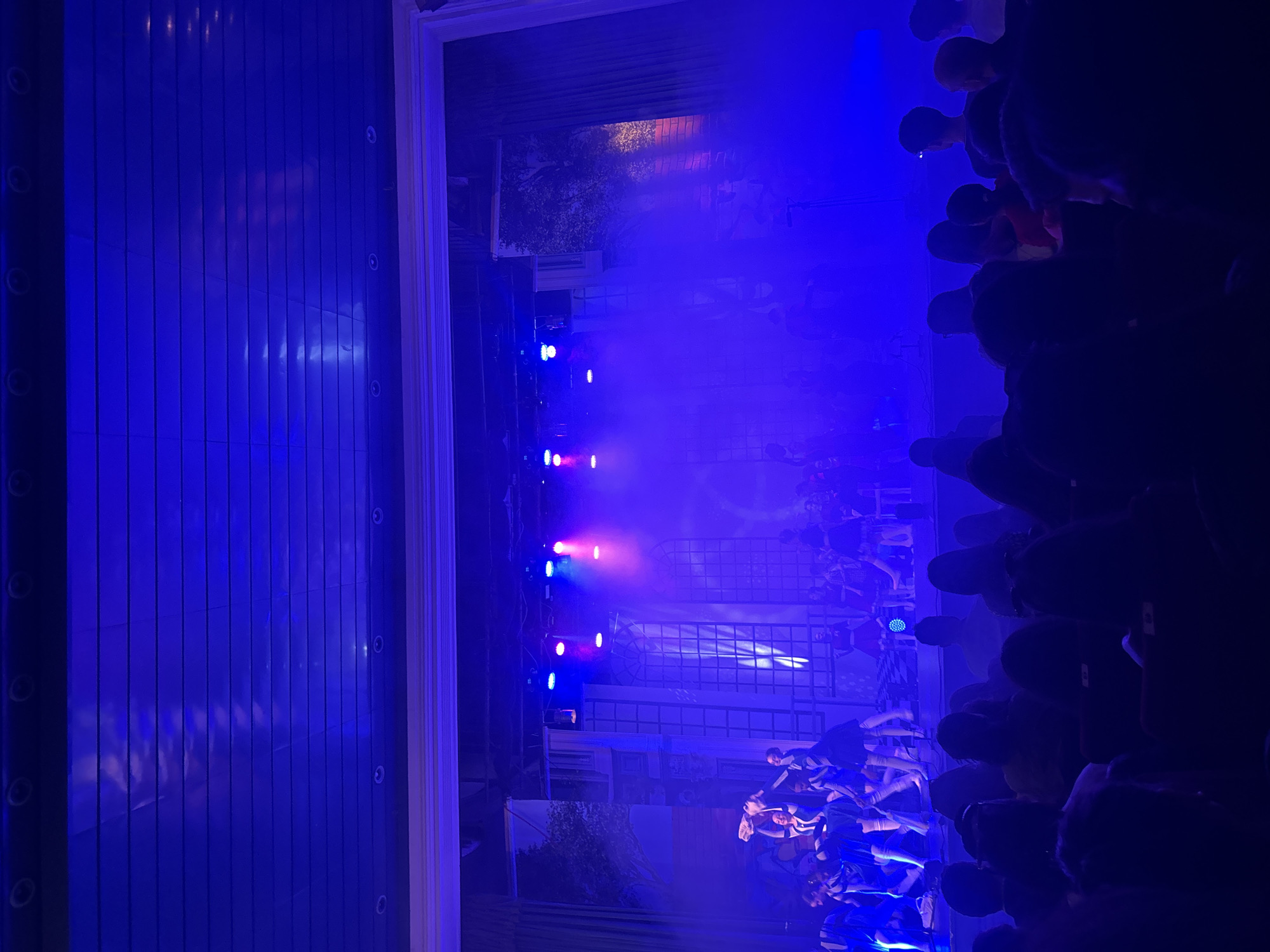 This image shows a crowd of people at a concert or event, viewed through a window. The scene is lit with blue and purple stage lights and fog. The crowd is dense, with many people holding up their phones. Outside of the window, there is a metallic exterior. The location is Sofia, Bulgaria.