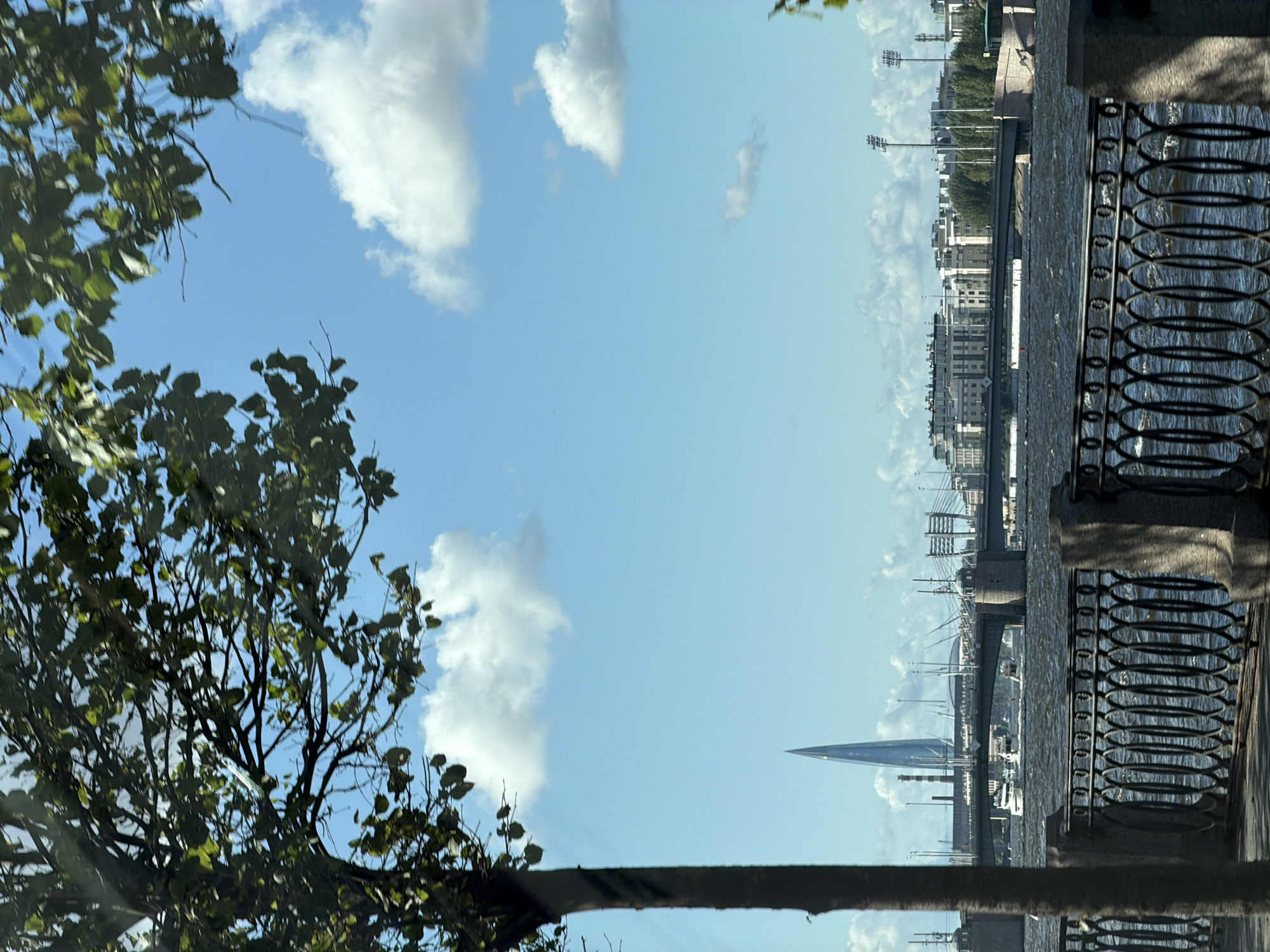 This is a photograph taken from the embankment of the Neva River in Saint Petersburg, Russia. The foreground features a dark, ornate metal fence with stone pillars, casting a shadow on the paved walkway. A large tree with dark bark and green leaves is positioned on the left side of the frame, obscuring part of the sky and city skyline.
Beyond the embankment, the Neva River flows with choppy water. Several boats, including a white yacht, are visible on the river. In the distance, a large bridge spans the river, with a modern skyscraper, the Lakhta Center, dominating the skyline behind it. The Lakhta Center is a very tall, slender skyscraper with a distinctive pointed spire. The city skyline also includes various buildings, some appearing to be residential or office blocks, and industrial structures. The sky is a clear blue with scattered white clouds. The overall scene suggests a daytime view of the city's waterfront.