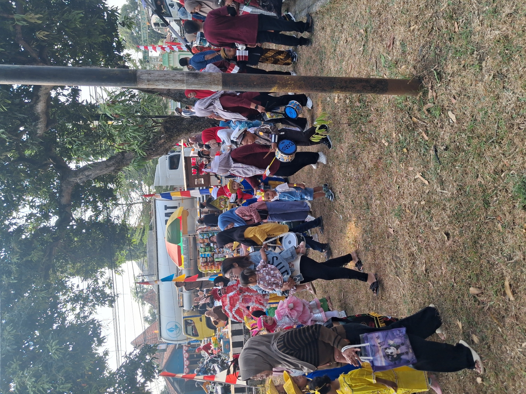 An outdoor event is taking place in Balapulang, Indonesia. A group of people, including children in costumes and adults, are walking on a grassy area. Some children are carrying drums, suggesting a parade or performance. In the background, there is a large truck, a colorful umbrella, and buildings. The scene appears festive, with flags and decorations visible. The ground is covered in dry grass and fallen leaves, and a tall pole stands prominently in the foreground. The overall atmosphere suggests a community gathering or celebration.
