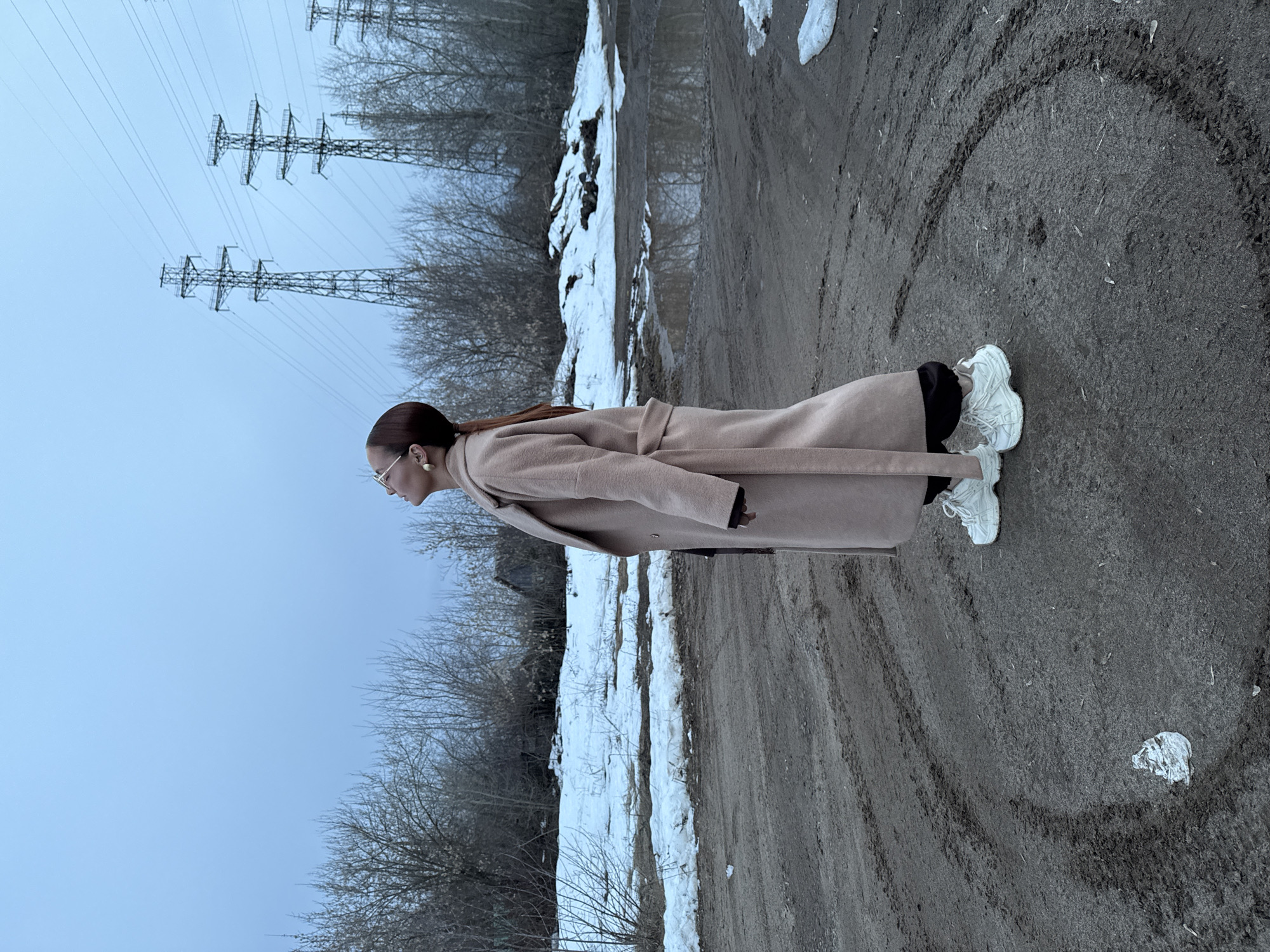 The image depicts a woman standing on a dirt road with tire tracks, possibly a rural area, in what appears to be the city of Chaikovsky, Russia. Traces of snow are present along the side of the road, next to a line of bare trees and power lines. The woman is wearing a light-colored coat and white sneakers. The ground shows circular markings in the dirt.