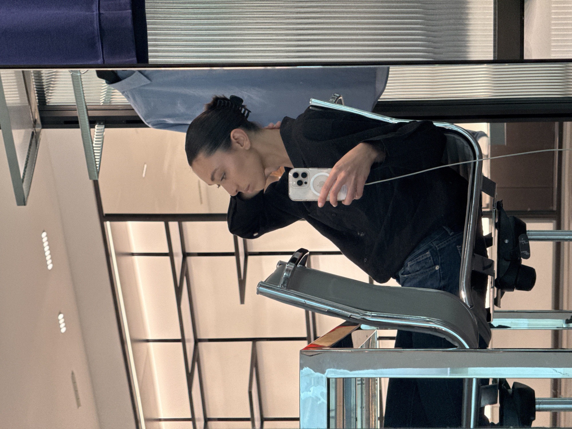 The image appears to be a mirrored reflection of a woman holding a smartphone in what seems to be a retail environment. The woman has dark hair pulled back with a hair clip, and is wearing a dark-colored top and jeans. She is seated in a modern, chrome-framed chair. The background shows elements of the store's interior design, including what looks like clothing racks or display shelving. There are also linear light fixtures on the ceiling, and what might be metal blinds. The location is likely within Milan, Italy, although specific details about the store are not visible.