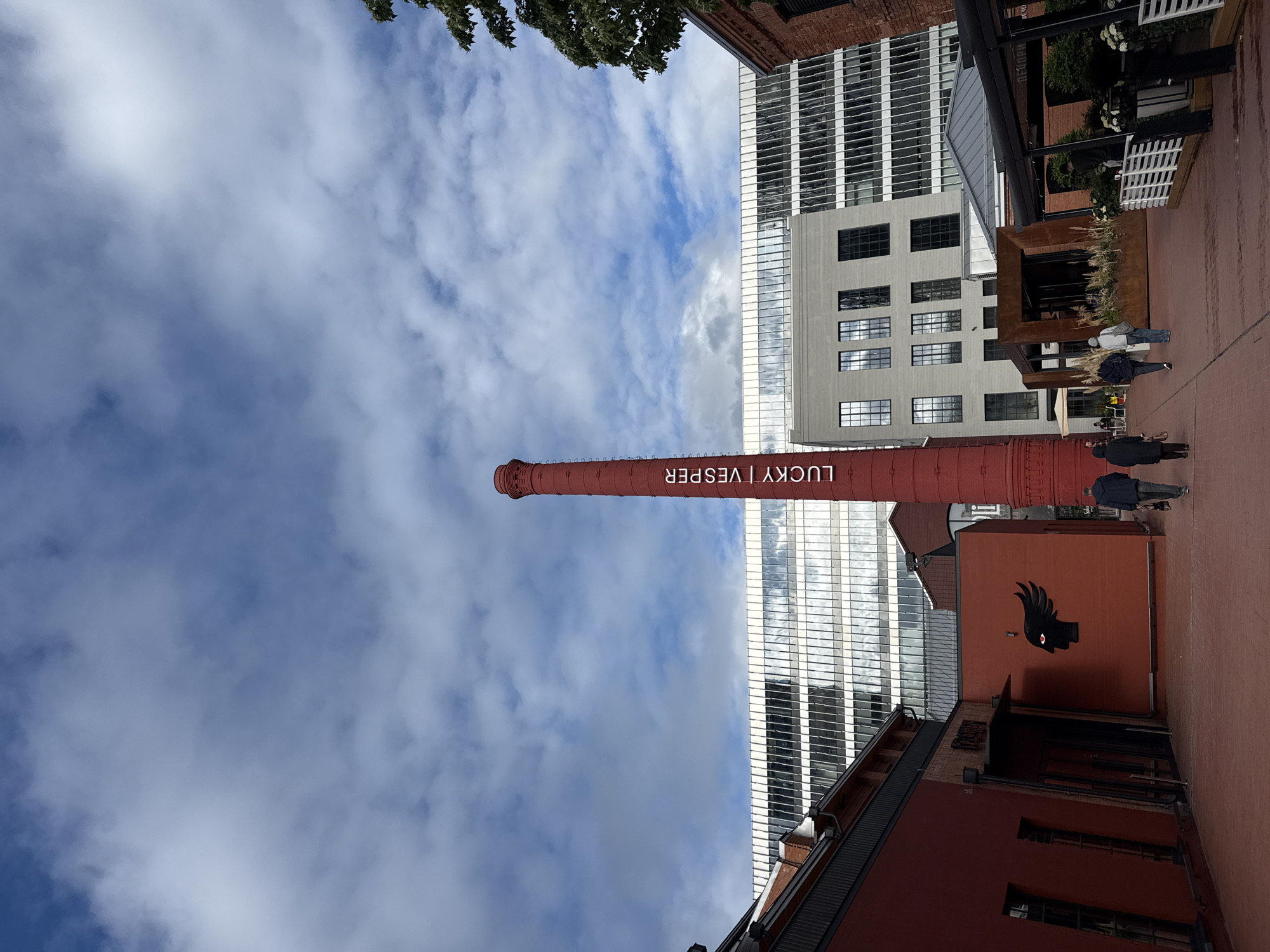 Captured from a low-angle perspective in Moscow, Russia, the photograph showcases a red brick chimney bearing the inscription "LUCKY | VESPER" against a backdrop of a partly cloudy sky. Adjacent to the chimney, a modern office building with a glass facade stands tall.
On the right side of the frame, people are visible, walking along a brick-paved ground. A brick building displays a prominent mural of a black silhouette of a head with a feathered crest, its eye marked with a red dot. The scene appears to be within a commercial or recreational area, combining historical industrial architecture with contemporary urban elements.