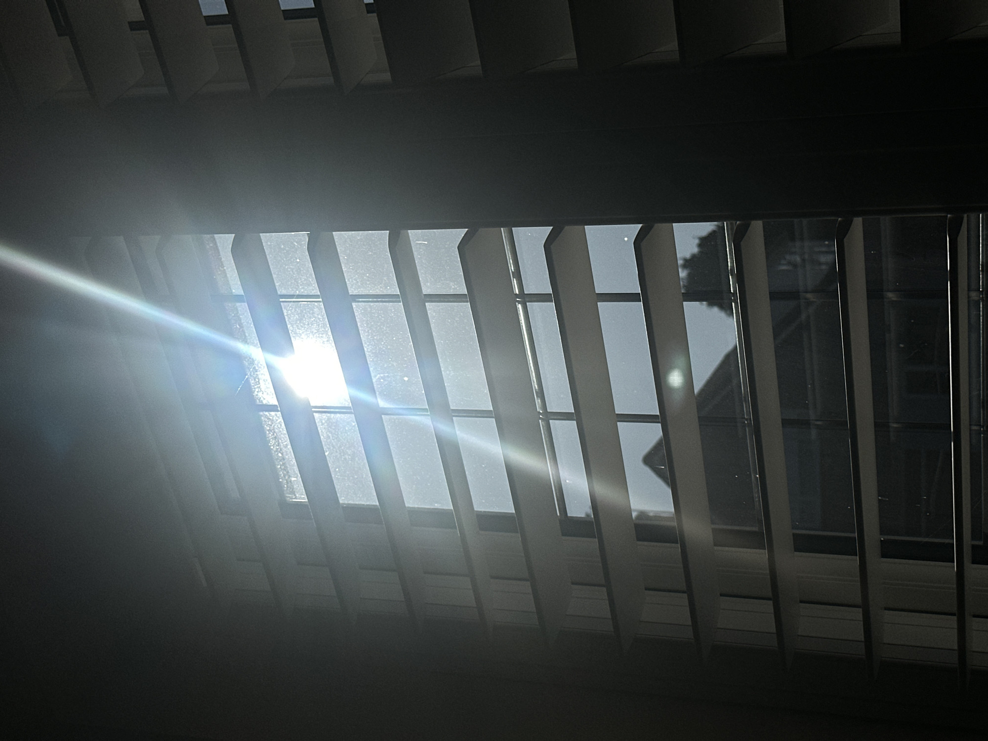 An indoor photograph from Wealden, United Kingdom, shows a window fitted with light-colored horizontal slatted blinds, viewed from an interior perspective. Intense sunlight penetrates the window, creating prominent lens flare and illuminating numerous dust particles on the glass and in the air. Through the gaps in the blinds, a bright sky is visible, along with the dark, silhouetted roofline of an external building and distant foliage. The interior surrounding the window is largely in shadow due to the strong backlighting. No individuals or specific actions are depicted.