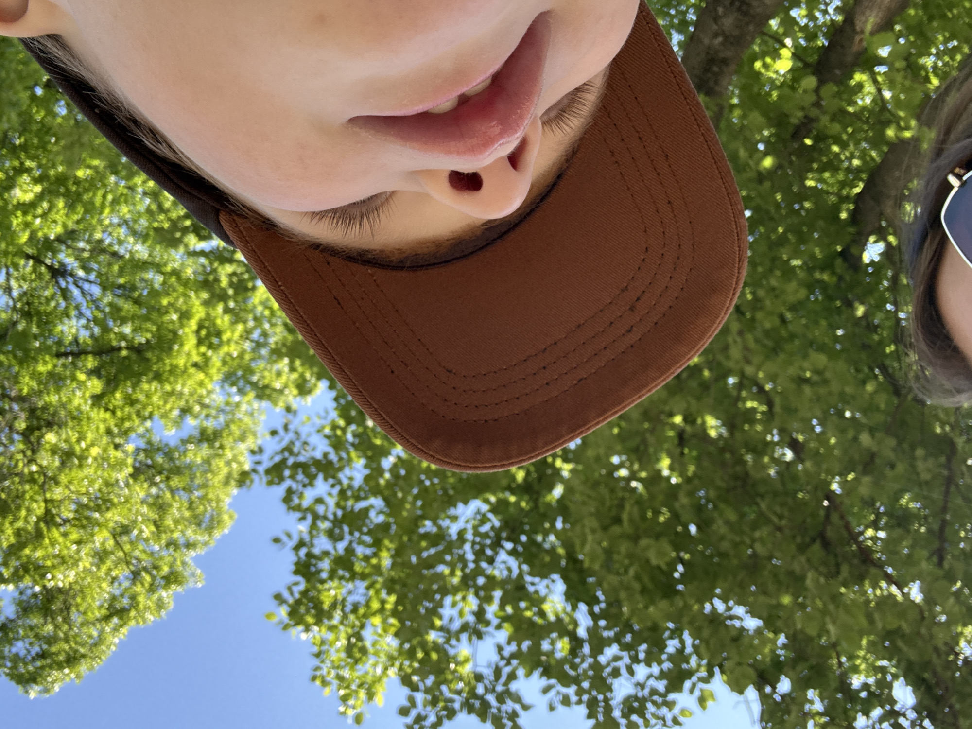 A low-angle, close-up shot shows a person wearing a brown baseball cap and looking up at the sky through the green leaves of a tree. The person's face is partially visible, with their mouth open and tongue slightly out. The background consists of a clear blue sky and lush green foliage. The scene is outdoors, likely in a park or natural setting, given the abundant tree cover. There is no indication of the city of Chaikovsky, Russia, within the image.