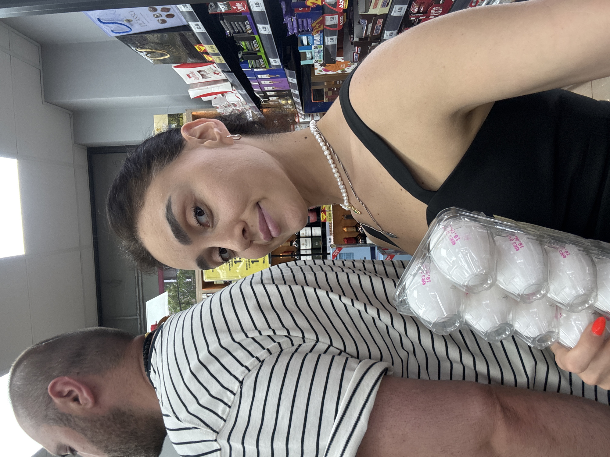 In this vertical, eye-level shot, a woman with dark hair pulled back and dark eyebrows is taking a selfie in a grocery store aisle. She is wearing a black tank top and a pearl necklace. She is holding a clear plastic container filled with small, white, individually wrapped items, possibly chocolates or candies, with pink text on them. To her right, partially visible, is a person wearing a black and white striped t-shirt, with their back to the camera. The background shows shelves stocked with various food products, including candy bars, protein bars, and other packaged goods, with price tags visible. Some visible text includes "PICASSO", "Raffaello", "Sante", and "PROTEIN 33%". The lighting suggests an indoor setting, likely a supermarket, during daytime hours. The woman's expression is neutral to slightly amused.