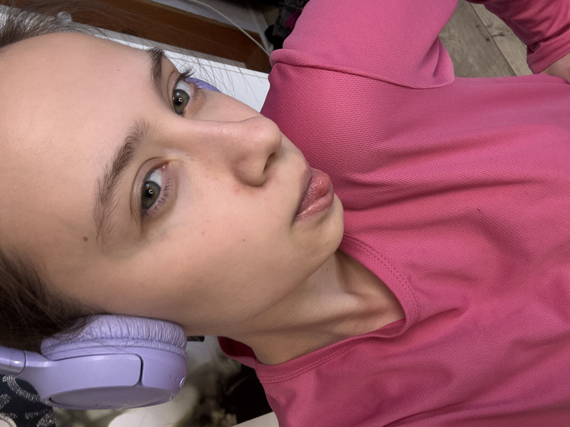 A young woman with fair skin, light brown hair, and green eyes is the subject of this close-up shot. She is wearing a pink long-sleeved shirt. Lavender headphones are visible next to her face. The background is blurred and contains a mixture of colors and indistinct shapes, possibly indicating an indoor setting with various objects. The orientation of the image suggests it was taken from a high angle, with the woman lying down or tilting her head towards the camera.
There is no indication of the image location being in the city of Прикамский, Russia.