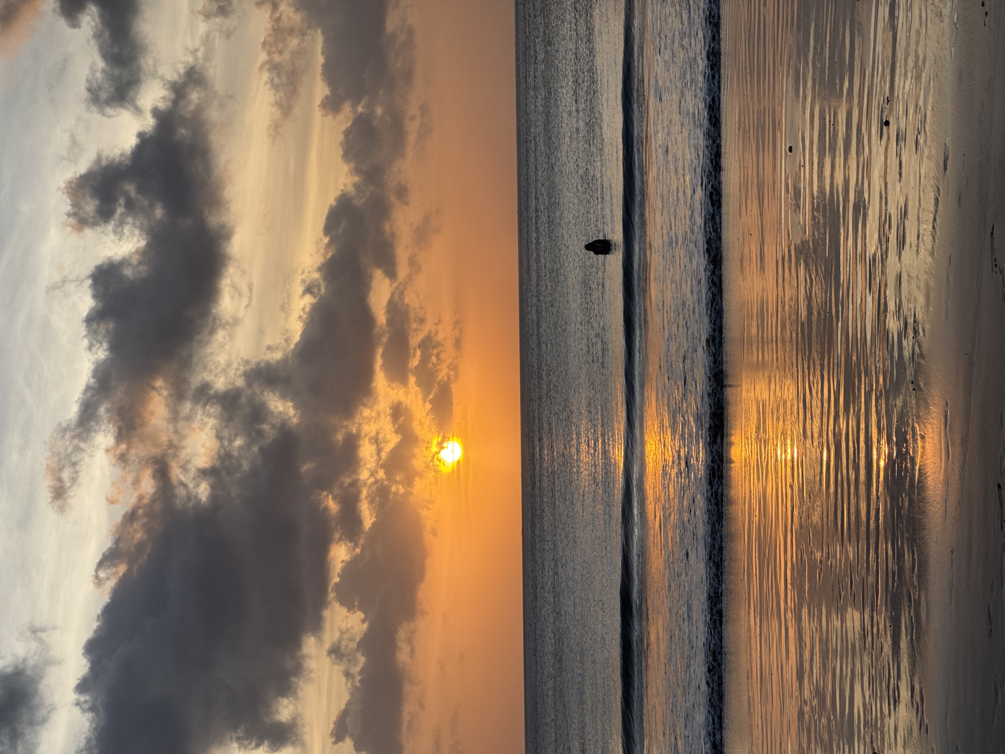 The image depicts a sunset scene at a beach in Kuta Selatan, Indonesia. On the left side of the image, the sky is filled with grey and orange clouds, with the sun visible near the horizon, casting an orange glow. The right side shows the ocean with small waves and a single person standing waist-deep in the water. The sand on the beach is visible in the lower-right corner, with sunlight reflecting on the wet surface.