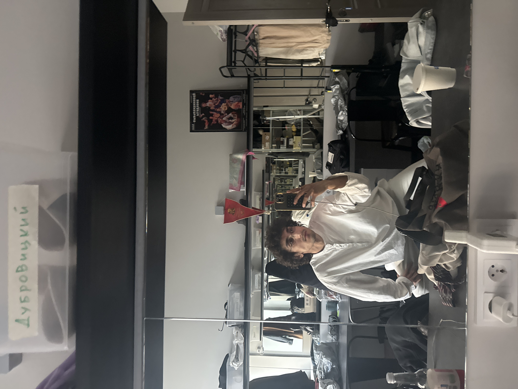 A man with curly dark hair, wearing a white long-sleeve shirt, is taking a selfie in a mirror. He is holding a phone in his right hand, and a small red flag is sticking out of the top of the phone. The background appears to be a cluttered dressing room or wardrobe area. There are shelves filled with various items, including clothes, accessories, and personal belongings. A poster is visible on the wall. The mirror reflects the room, providing a view of the overall layout. The name "Дубровицкий" is visible on a tag attached to something near the mirror's edge. An electrical outlet is mounted on the wall near the mirror. Given the flag, the man, and the name, the image is likely taken somewhere in Russia, possibly Moscow.