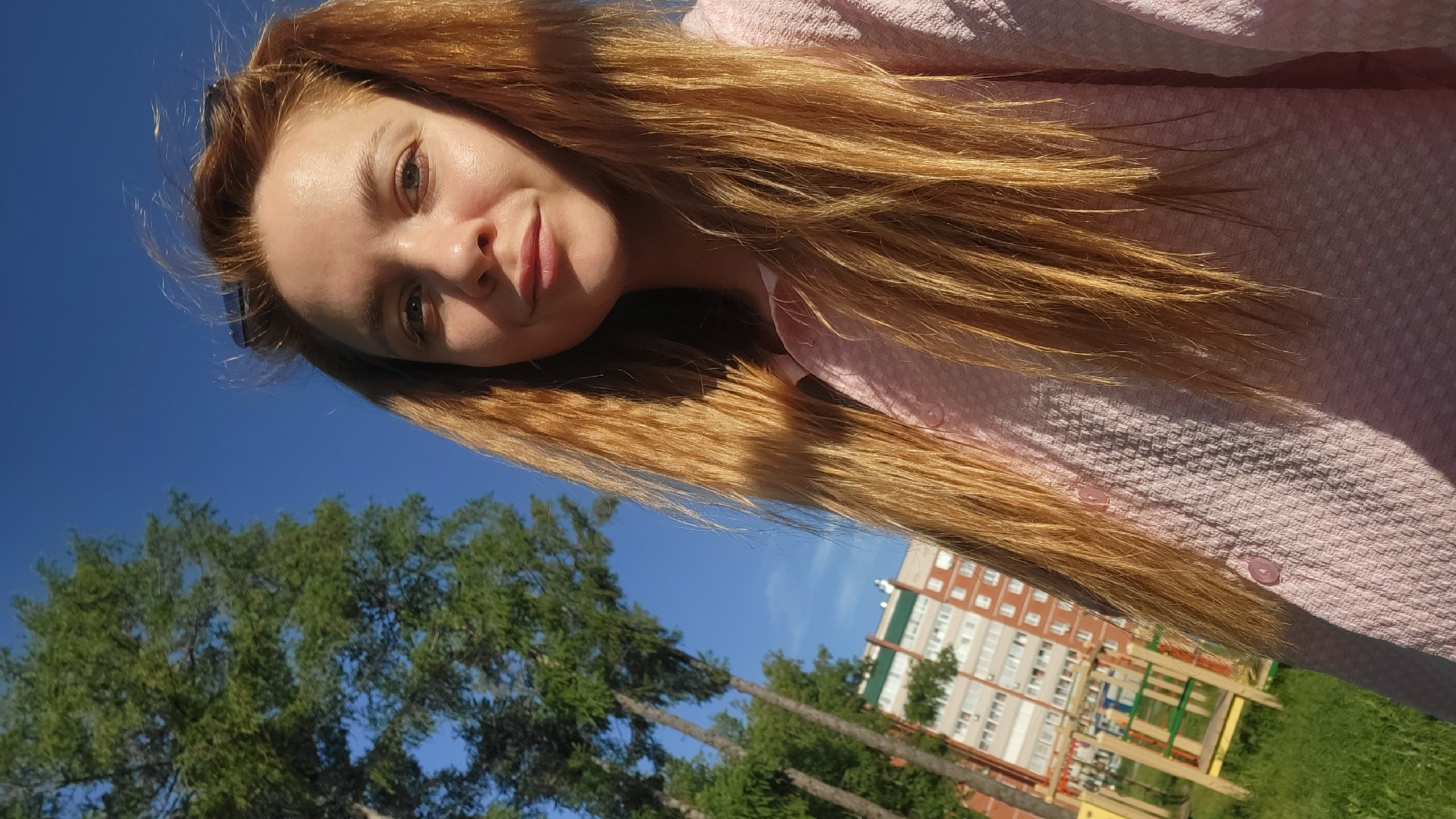 A selfie photograph shows a fair-skinned woman with long, light brown hair wearing a pink, textured button-down shirt. The image is taken from a low angle, slightly tilted to the left. The woman's face is partially in shadow, but her features are visible, including her eyes, nose, and a slight smile.
The background includes a clear, blue sky at the top and green foliage of trees in the lower-left corner. A multi-story brick building with white window frames is visible in the lower-right, along with a wooden playground structure in front of it. The location is in the city of Chaikovsky, Russia, as the building's architectural style and the general environment are consistent with that region.