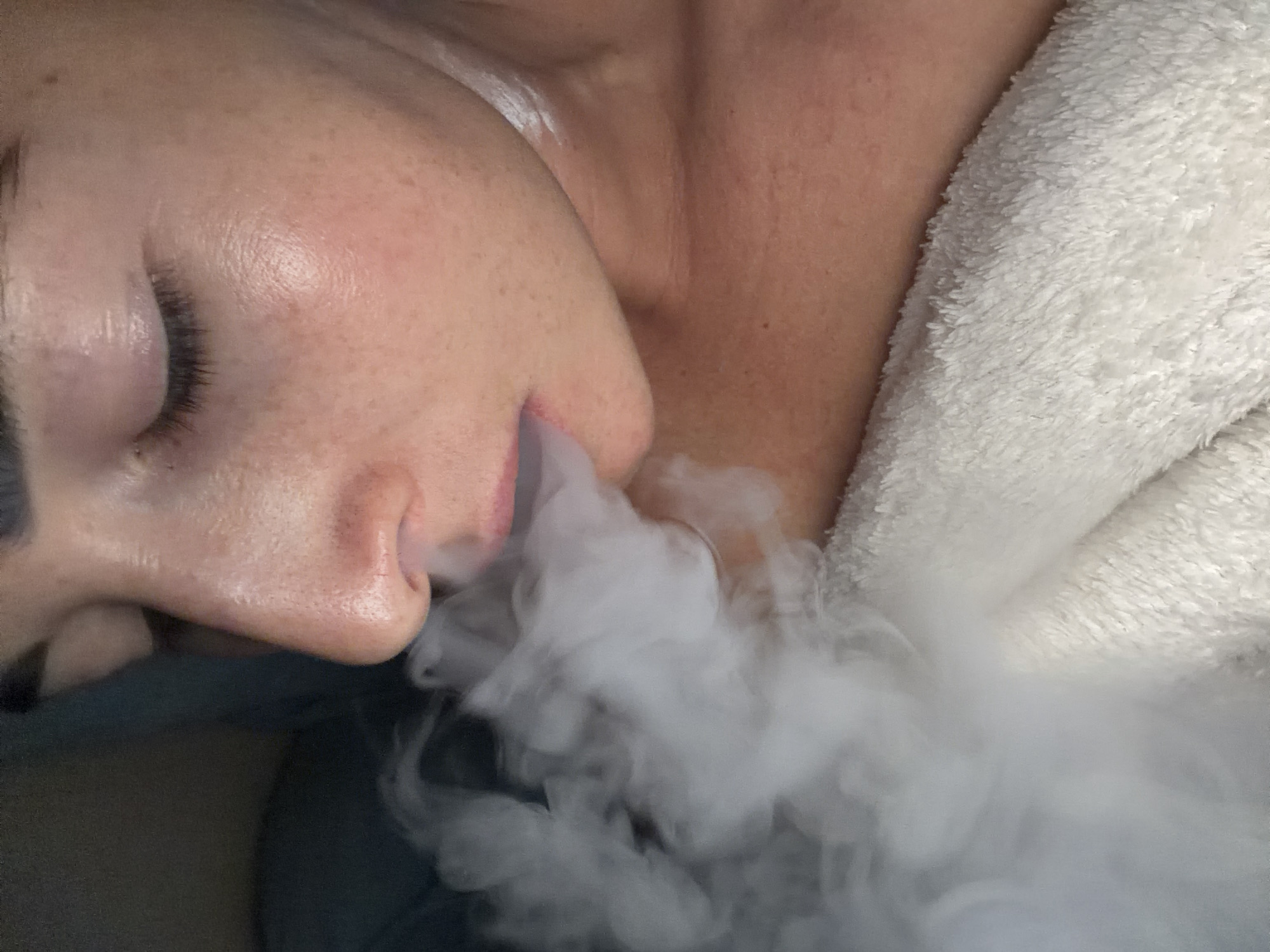 A close-up shot shows a person lying on their side, exhaling a large cloud of white smoke. The person has fair skin, freckles, and closed eyes with dark eyelashes. The smoke obscures part of their face and chest. A fluffy white towel is draped around the person's shoulder and chest. The location is Unknown, Russia.