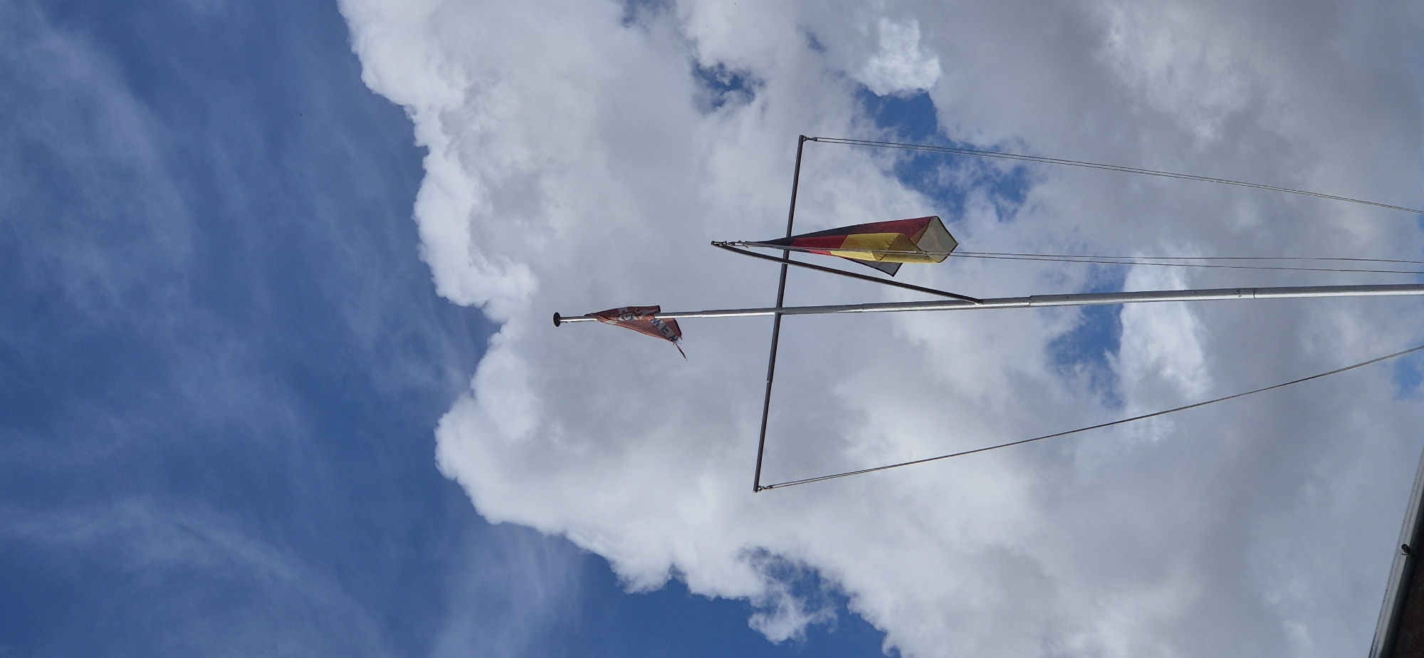 The image shows a flagpole against a cloudy blue sky. A German flag, featuring black, red, and gold horizontal stripes, hangs from the pole. There is also a smaller orange flag with some markings, possibly a crest or logo, flying slightly higher on the pole. The flagpole appears to be made of metal and has some rigging attached. The background consists of large, white cumulus clouds with patches of blue sky visible in between. The lower part of the image shows a glimpse of a dark-colored roof or structure, suggesting the flagpole is located on a building or vessel. The scene is devoid of people or any discernable activity. The context provided suggests the location is the city of Mainz, Germany, though this cannot be visually confirmed from the image itself.