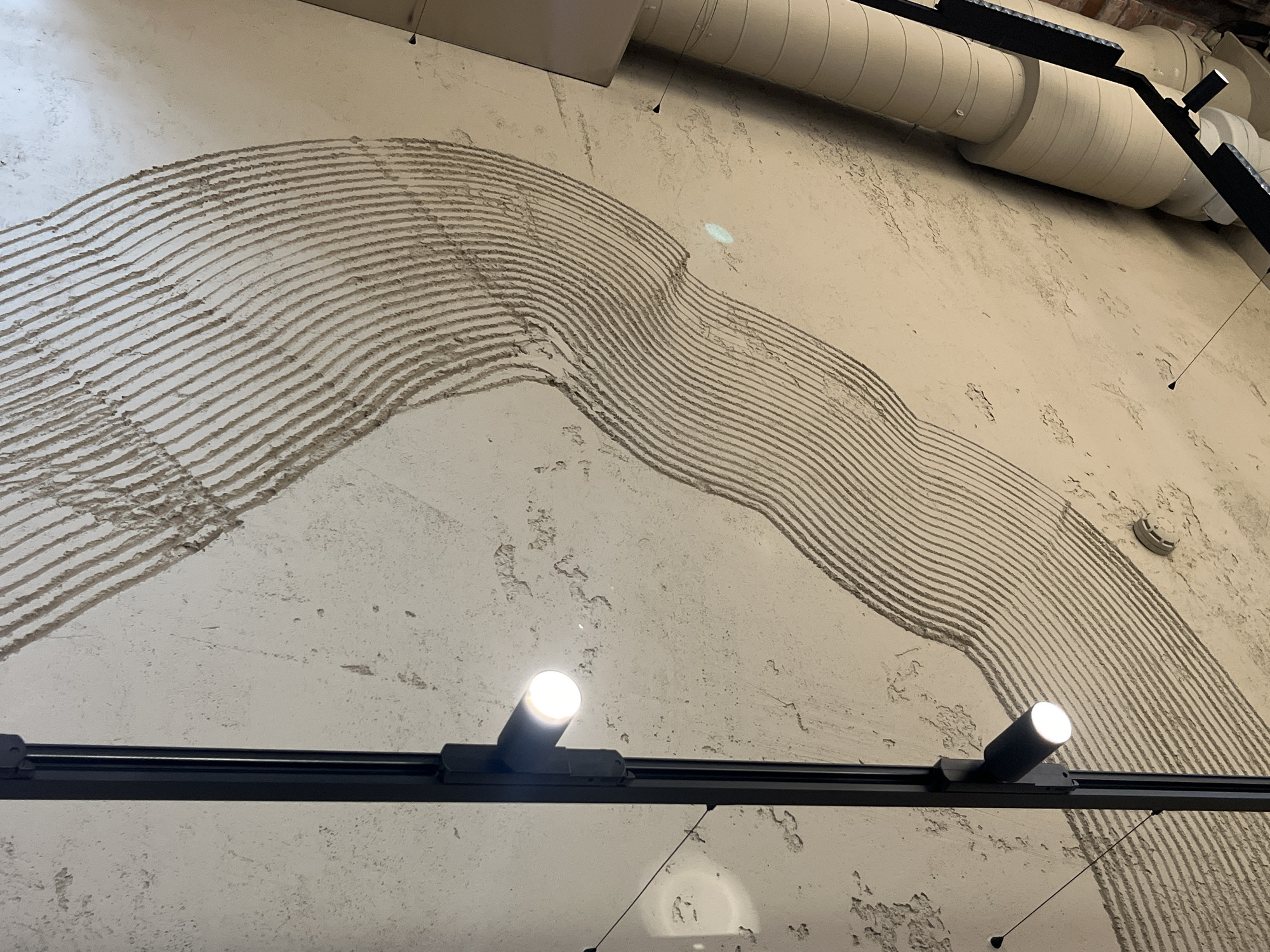 This image captures a section of a textured ceiling in Svetlogorsk, Russia. The ceiling appears to be made of concrete or a similar rough material, with a patterned texture created by parallel, wavy indentations. A black track lighting system is mounted vertically on the left side of the frame, with two spotlights visible. One spotlight is illuminated, casting a bright light. To the right, a large beige pipe or conduit runs diagonally across the upper right portion of the image, appearing to be part of an industrial or architectural feature. The lighting suggests an indoor environment, possibly a public space or a commercial building, due to the track lighting and the exposed pipe. The overall impression is one of modern or industrial interior design.