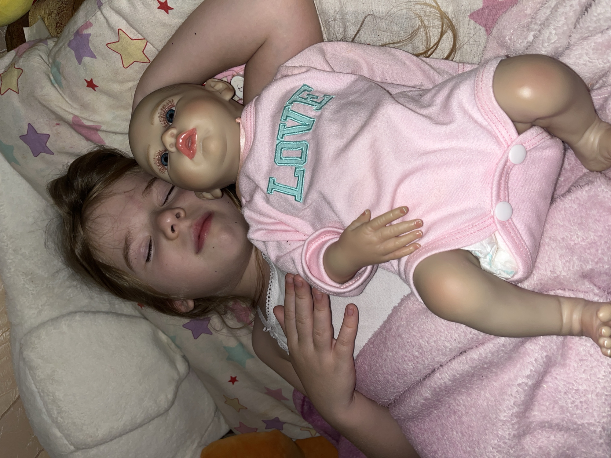 An indoor scene depicts a young child and a doll lying side-by-side on a bed, appearing to be asleep. The child, with light brown hair, is positioned on the left, with eyes closed and mouth slightly ajar. They wear a white camisole. Their left arm rests over the doll, while their right arm is under a pink, textured blanket. The doll, a reborn-style figure, has fair skin, blue eyes, painted pink lips, and prominent reddish-gold eyelashes. It wears a pink long-sleeved onesie with "LOVE" embroidered in teal letters on the chest, and a white diaper is visible underneath. The doll's small hands have visible painted fingernails. The subjects are resting on a light-colored sheet or pillowcase adorned with pastel-colored stars (yellow, purple, light blue, pink, and red). A white, textured pillow supports the child's head. The lighting is dim, suggesting a nighttime or low-light setting. The specific geographical location, such as Sochi, Russia, cannot be determined from the visual information presented in the image.