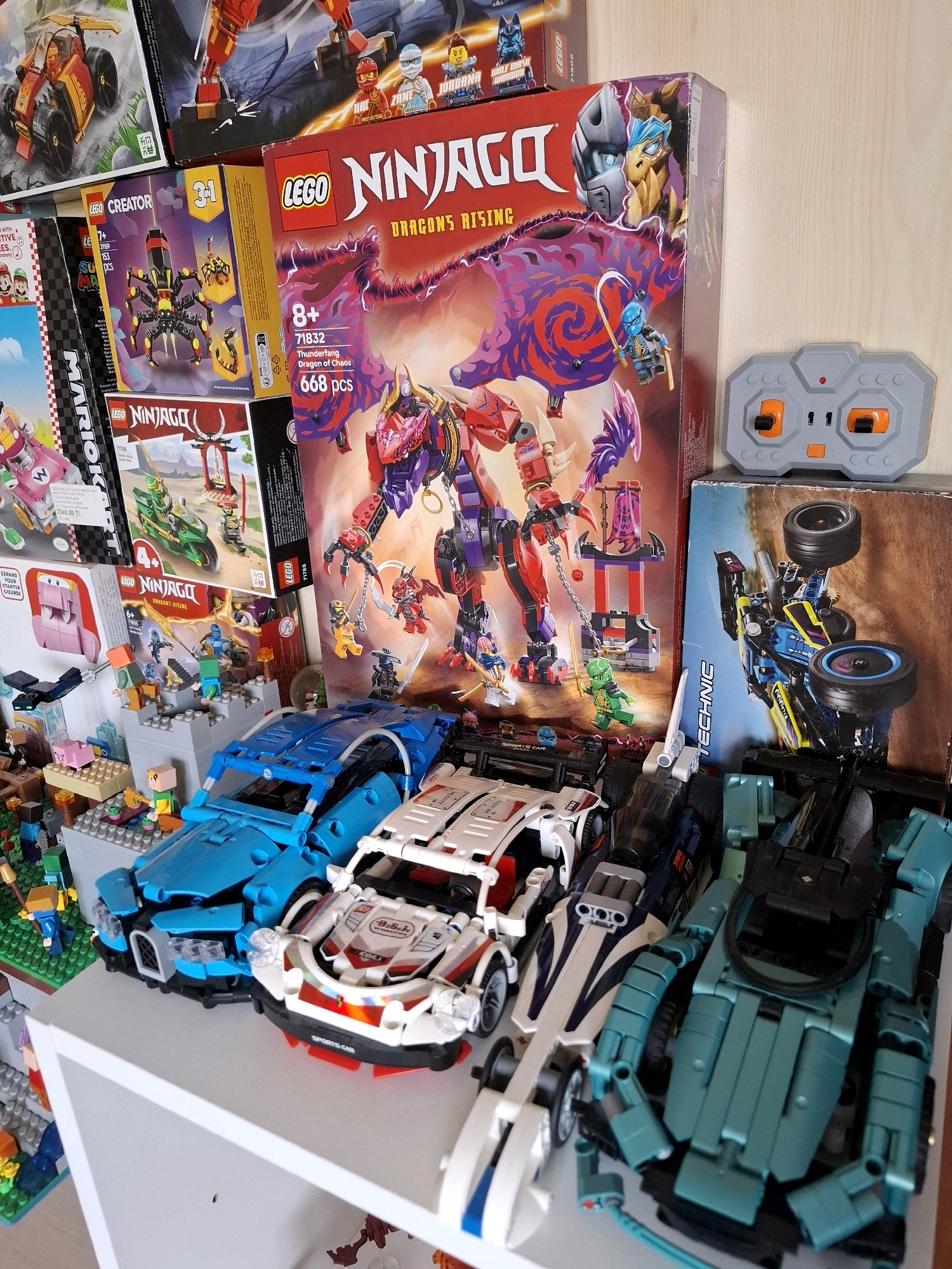 The image displays a collection of Lego sets, predominantly from the Ninjago and Creator series, arranged on shelves against a plain wall. The Lego sets feature various vehicles, characters, and buildings. In the foreground, several Lego cars are visible, including a blue sports car, a white and red sports car, and a teal-colored vehicle. Behind the cars, more Lego construction sets and boxes are stacked, including a large Ninjago box with a dragon illustration and a remote control for a Lego set. The scene appears to be a domestic setting, possibly a child's room or a collector's display. There is no indication of the city of Tyumen, Russia, or any specific location. The lighting suggests an indoor environment.