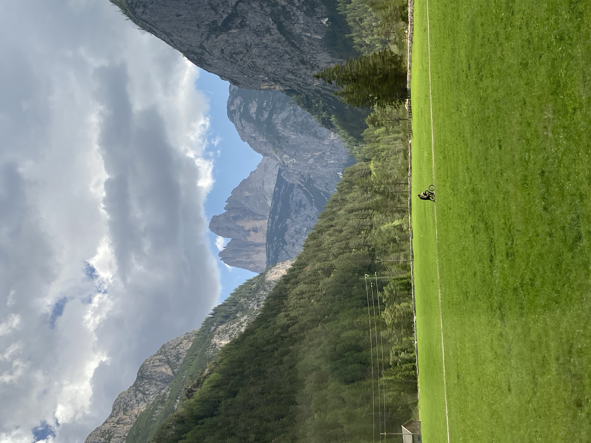 The image depicts a scenic mountain landscape in the vicinity of Toblach (Dobbiaco), Italy. In the foreground, a vast expanse of lush green grass covers a meadow. A single person is seen riding a bicycle on a white path that traverses the field. To the left, a small wooden building, possibly a shed or cabin, is visible at the edge of the meadow, near a treeline. The middle ground is dominated by a dense forest of green coniferous trees that climb the slopes of the mountains. In the background, a majestic mountain range rises, characterized by rocky peaks and steep inclines. Notably, the iconic Tre Cime di Lavaredo (Three Peaks of Lavaredo) are visible in the distance, their distinct sharp summits piercing the sky. The sky above is a mix of dramatic clouds and patches of blue, suggesting dynamic weather conditions. Power lines with poles are present across the landscape. The overall impression is one of natural beauty and outdoor recreation in a mountainous region.