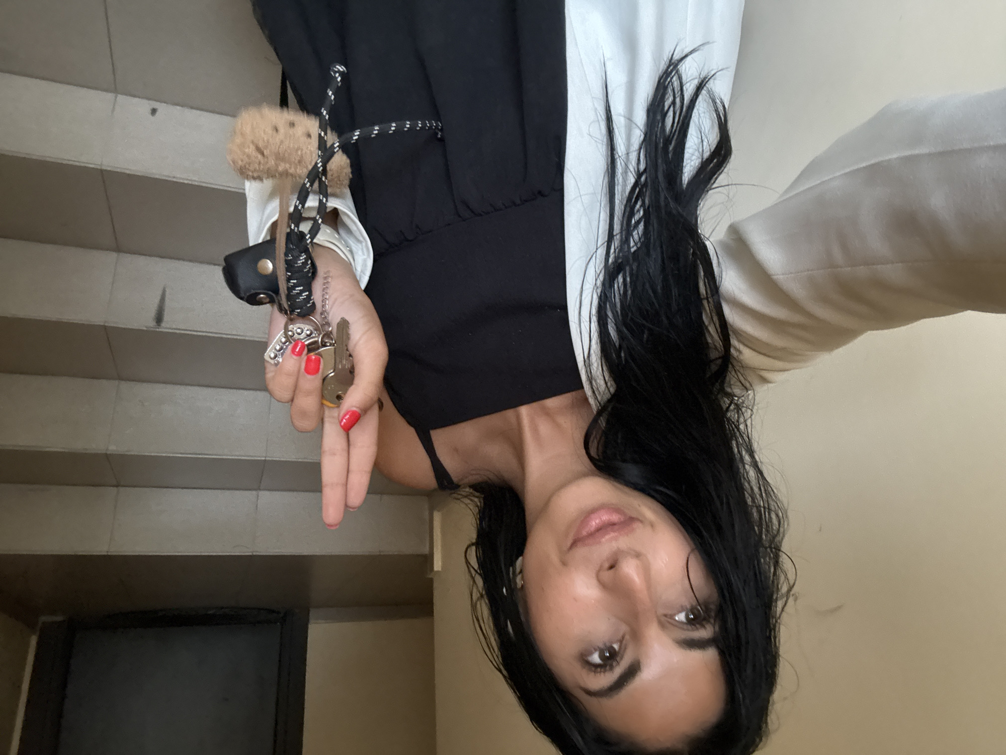 A woman with long, dark hair, wearing a black dress and a light-colored jacket, stands on a tiled staircase. She holds a set of keys in her right hand, displaying them to the camera. The background shows a dark door and the continuation of the tiled steps. The scene appears to be an interior staircase within a building, possibly an apartment complex or office building. There is no specific visual information in the image to link it to the city of Plovdiv, Bulgaria.