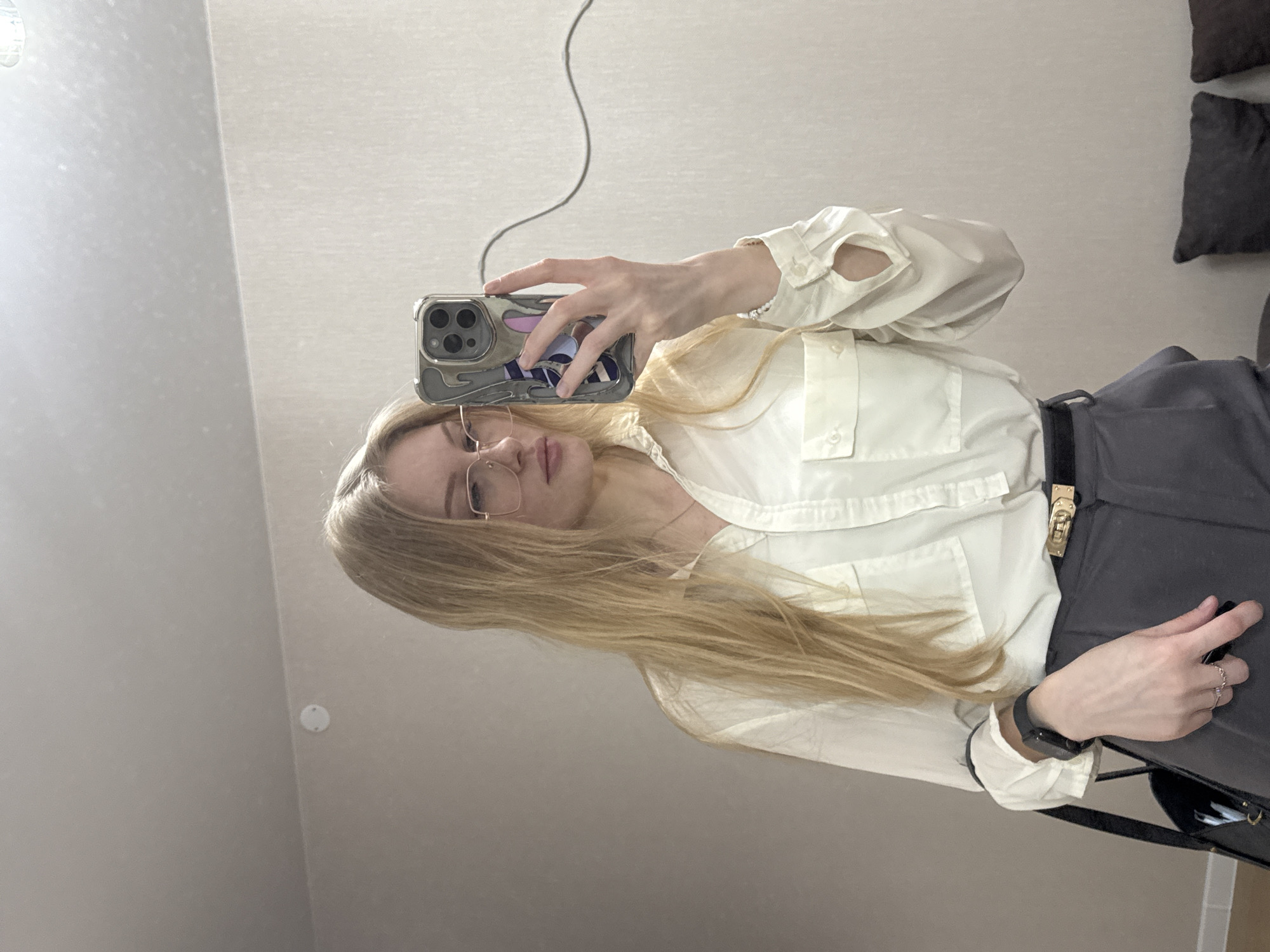 A mirror selfie captures a young woman with long, straight blonde hair, wearing clear-framed glasses, a cream or off-white long-sleeved button-up shirt with two breast pockets, and dark grey trousers cinched with a black belt featuring a gold-toned buckle. She holds a silver smartphone, featuring a clear, patterned case, in her right hand to take the photo. A black smartwatch or fitness tracker is visible on her left wrist, along with a silver-colored ring on her ring finger. A dark bag or purse is partially visible beside her left hand. Her expression is neutral.
The indoor setting features light-colored, possibly textured walls and a light ceiling, where a white circular smoke detector is visible. Two dark grey throw pillows rest on a surface in the upper right background. A dark, wavy cord hangs down in the reflection of the mirror. The image appears to be taken during the daytime, though no specific time or weather details are evident. There is no visible text in the image. This scene is contextualized in Perm, Russia.