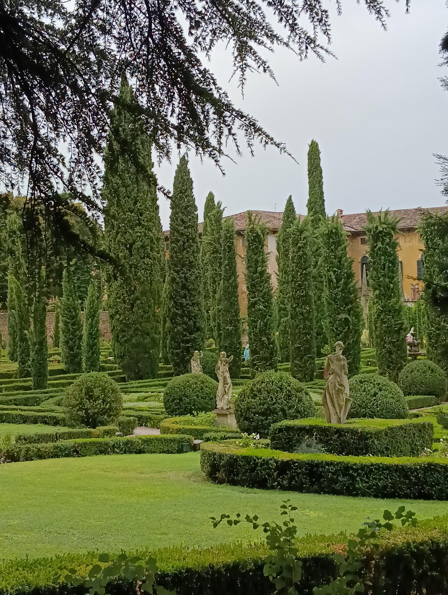 The image depicts a formal Italian garden, likely at Giardino Giusti in Verona, Italy, characterized by meticulously manicured hedges, topiaries, and tall cypress trees. Stone statues are strategically placed throughout the garden, one of which is prominently visible in the foreground, depicting a draped female figure. In the background, a large building with a terracotta roof and yellow facade suggests the presence of a villa or palace. The sky is overcast, casting a soft, diffused light over the scene. Two figures, one in blue and another in pink, are faintly visible walking in the middle ground, adding a sense of scale and life to the otherwise static landscape. The garden appears to be well-maintained and offers a sense of classical beauty and tranquility.
