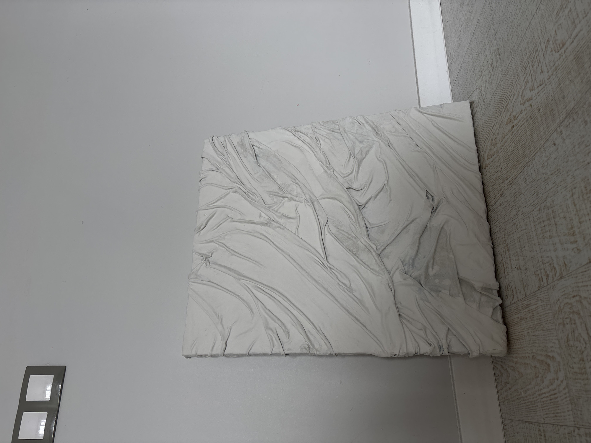 This media file presents an interior scene featuring a prominent, textured, rectangular object. The object, appearing to be a relief sculpture or a heavily textured canvas, is light grey to off-white and measures roughly half the width of the frame. Its surface is characterized by complex, three-dimensional folds and drapes, resembling rumpled fabric such as bedsheets or crumpled cloth. These textures create varied shadows and highlights, emphasizing the object's depth.
The object is positioned against a plain, light grey wall, which occupies the majority of the background. In the lower left corner of the wall, a dark grey or black electrical outlet or light switch with two rectangular reflective sections is visible. To the right of the textured object, a light-colored wooden or laminate floor is depicted, showing a distinct grain pattern that runs diagonally. A thin, white vertical trim separates the wall and the floor in the upper right.
The scene is static, with no activity or events taking place. The lighting is even, suggesting ambient indoor illumination rather than direct sunlight. The overall color scheme is neutral, dominated by shades of grey and white, with the warm, light brown tones of the floor providing a contrast. A small, faint pink or purple speck is visible on the plain wall in some detailed views. No people or visible text are present in the image.