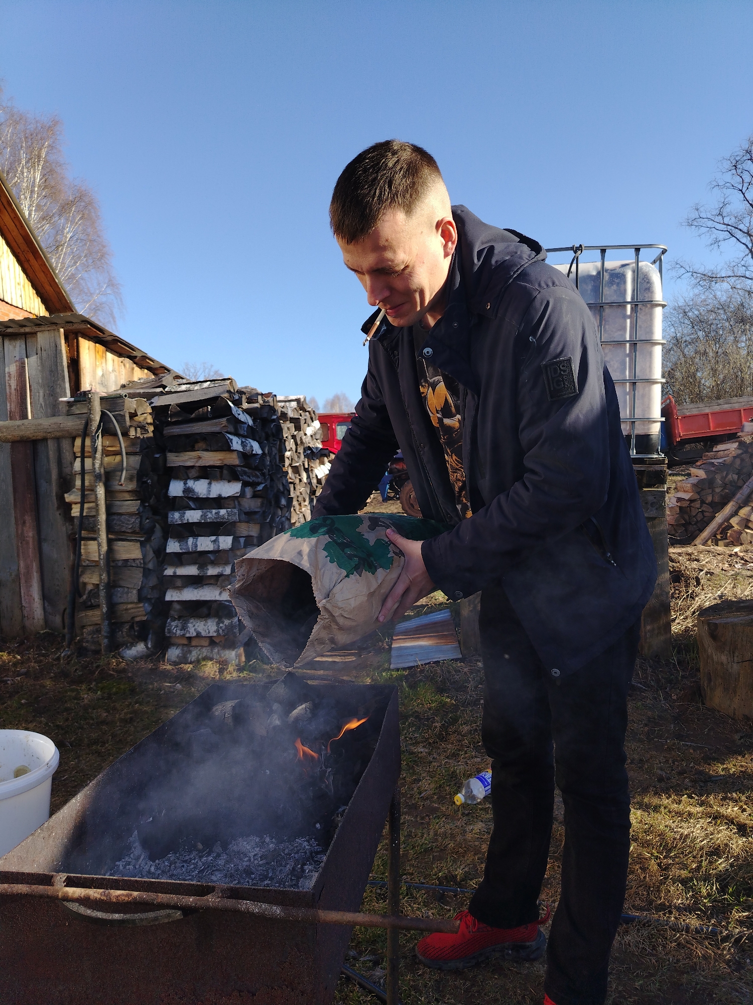 A man stands outside in a rural setting, possibly in the city of Nizhnyaya Gar', Russia, based on the prompt. He is wearing a dark jacket, black pants, and red sneakers. He is holding a paper bag and pouring its contents into a rusty metal grill. Smoke and flames are visible inside the grill, suggesting it contains burning materials, likely charcoal. The man has a cigarette in his mouth. Behind the man, there is a wooden structure, a stack of firewood, and a large white plastic container with a metal frame. A discarded plastic bottle is on the ground next to the man. The weather appears sunny with a clear blue sky.