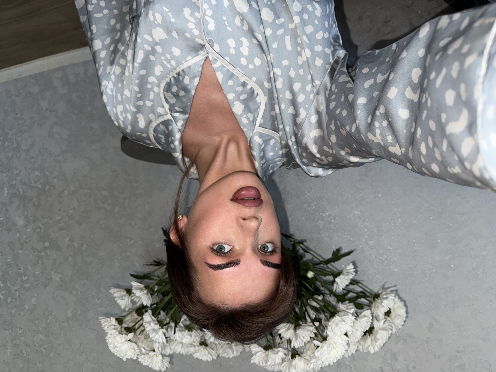 The image shows a woman lying on a light gray textured surface with a bouquet of white chrysanthemums beneath her head. She is wearing a light blue pajama top with white spots and white piping. Her hair is brown, and she has on light pink lipstick. She is looking directly at the camera. The image was taken in Лобаново, Russia.