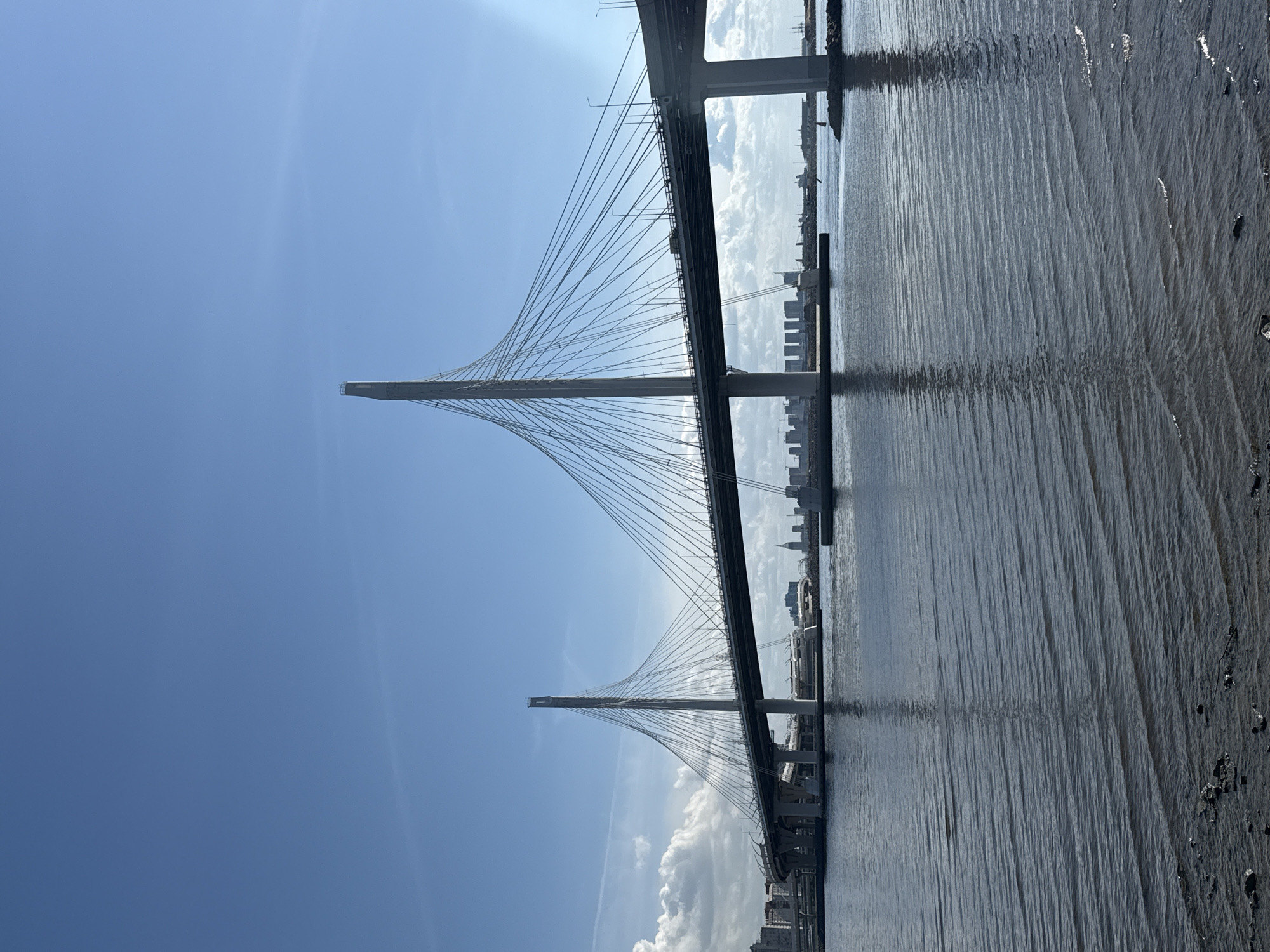 The image depicts the Saint Petersburg cable-stayed bridge in Russia. The bridge is viewed from a low angle, emphasizing its towers and cable system. The sky is blue with some clouds. The water reflects the bridge structure and the skyline of Saint Petersburg.