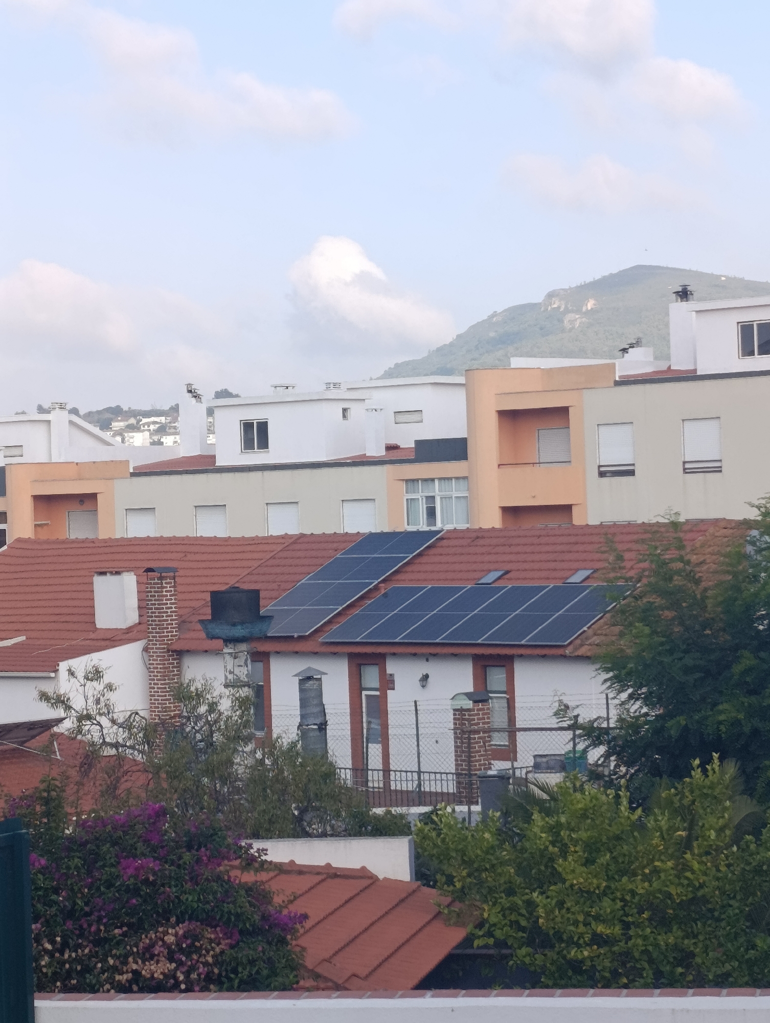 The image displays a view of residential buildings in Setúbal, Portugal, under a cloudy sky. The foreground features red-tiled rooftops, some equipped with solar panels. A prominent brick chimney stands on one of the roofs, with a black cylindrical object on top. Lush green trees and purple flowering bushes are also visible in the lower part of the frame, partially obscuring some buildings. In the mid-ground, several multi-story buildings with white, beige, and peach facades are present, many with white window shutters. The background is dominated by a large, green, mountainous landscape. The overall scene depicts a residential area with a mix of traditional and modern architecture, set against a natural backdrop.