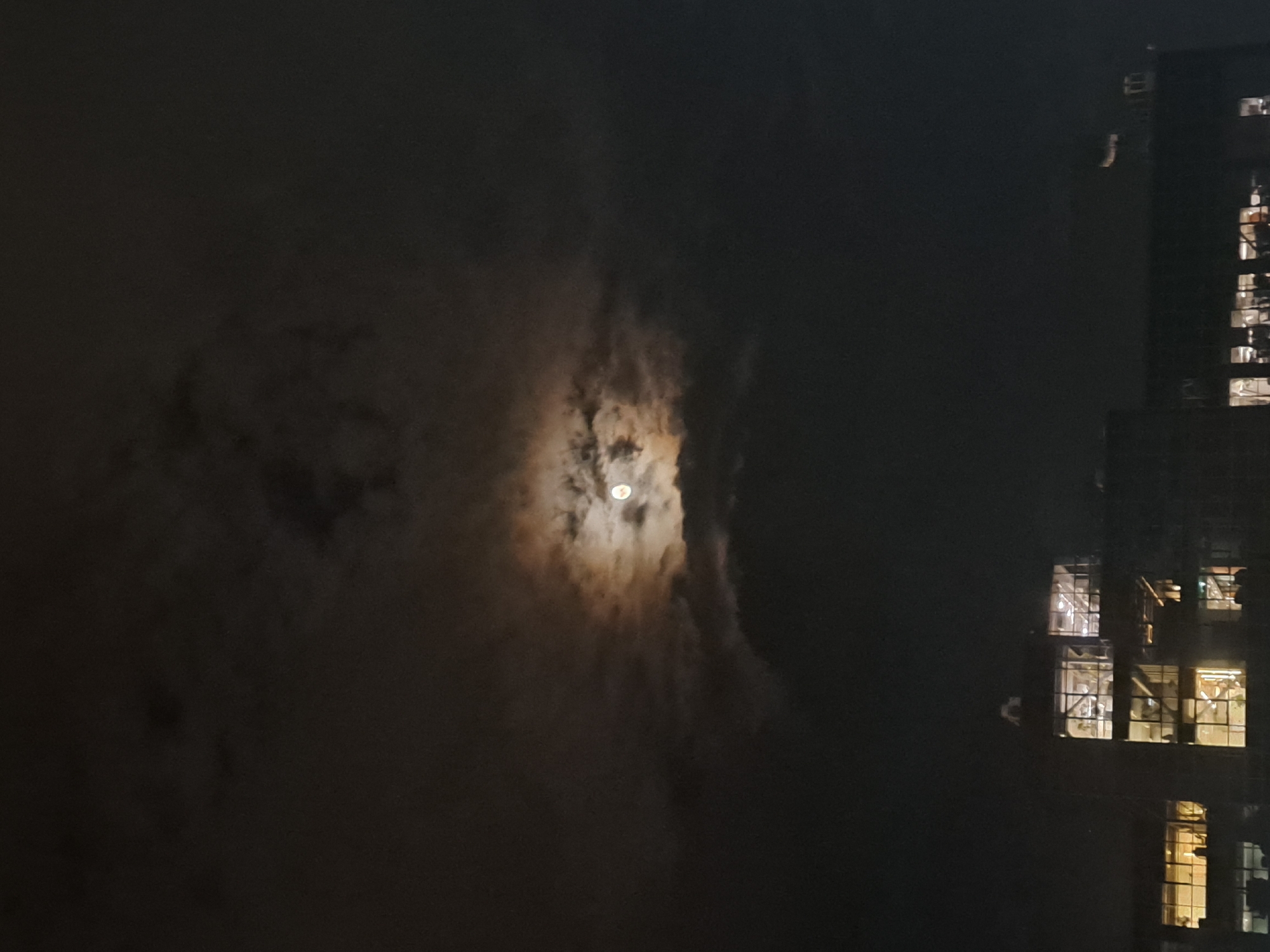 The image depicts a nighttime urban scene in Yerevan, Armenia, focusing on a luminous moon partially veiled by clouds, and a well-lit building. The sky is dark, with a significant portion covered by dense, dark clouds. Through a dramatic vertical opening in these clouds, the full moon shines brightly, casting a soft, warm glow that illuminates the edges of the surrounding cloud formations, creating a striking contrast against the darker sky.
To the right of the frame, a tall, multi-story building stands against the dark night. It appears to be a modern structure, possibly industrial or commercial, given the visible grid-like patterns within its windows. Many of the building's windows are brightly illuminated from within, emitting a warm, yellow-white light that sharply contrasts with the subtle moonlight. The internal lighting reveals structural elements or possibly scaffolding within several window sections, suggesting either ongoing construction or a functional, perhaps industrial, interior.
The scene conveys a serene yet dynamic urban night, where the natural light of the moon interplays with the artificial glow emanating from human architecture. No discernible text is visible within the image.