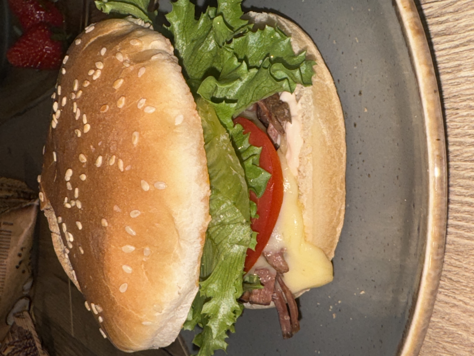 A close-up, eye-level shot captures a substantial sandwich presented on a rustic, dark-colored ceramic plate in an indoor setting, likely in Krasnoyarsk, Russia.
The primary subject is a hearty sandwich or burger, featuring a golden-brown sesame seed bun that is split open, revealing its various fillings. Inside, fresh, vibrant green leaf lettuce is layered prominently, followed by a bright red slice of tomato. Beneath these, a generous amount of melted yellow cheese cascades over thinly sliced, cooked brown meat, which appears to be roast beef or a similar cut. A creamy white sauce is also visible, enhancing the sandwich's composition.
The sandwich rests on a speckled dark grey or blue-grey plate with a distinctive light brown rim. In the background, to the left of the sandwich, two ripe red strawberries are partially visible, suggesting they accompany the meal. A fragment of a light brown paper bag or packaging with faint, unreadable text is also present in the bottom-left background. The entire meal is set on a warm-toned wooden tabletop, which is visible on the right side and in the bottom-left corner of the frame.
The lighting in the image is somewhat dim, implying an indoor environment, potentially during the evening, with artificial light illuminating the food. There are no individuals present, and the image functions as a still life, showcasing a prepared meal ready for consumption.