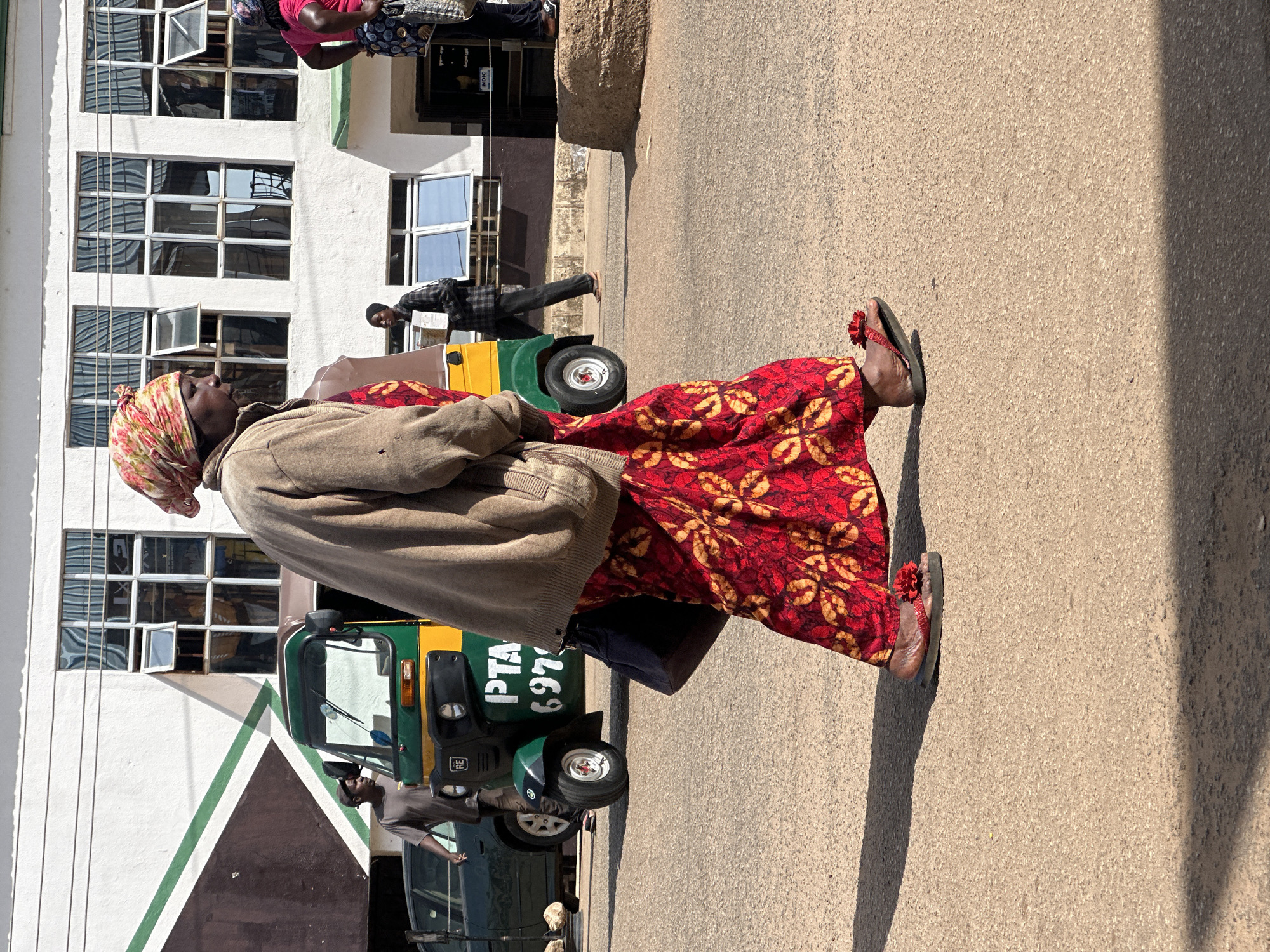 In Bukuru, Nigeria, a woman wearing a red and yellow patterned headscarf, a tan cardigan, and a long red patterned dress walks across a paved street. She is carrying a dark bag in her left hand. Beside her, a green and yellow auto-rickshaw is parked. In the background, a man in a gray shirt stands near the auto-rickshaw. Further back, a white building with multiple windows is visible. Another person in a pink shirt and patterned skirt is seen on the right side of the image, facing away from the camera. The scene appears to be a street in a commercial area, with people and vehicles present.