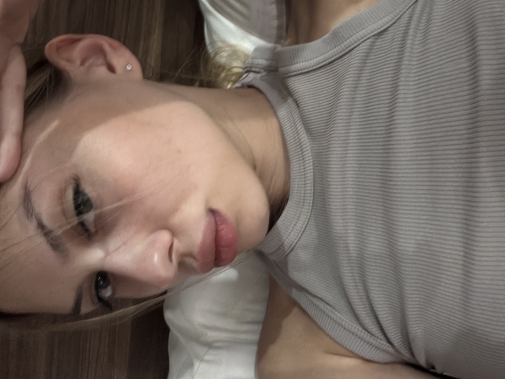 A close-up, upper-body shot features a young woman with blonde hair and light skin. She is wearing a grey ribbed tank top and a small stud earring in her left ear. Her hand is resting on her forehead. The background is out of focus, appearing to be a bed with white pillows and a wooden headboard. The lighting is soft, with a slight shadow cast across her face. The image is undated and the location in Da Nang, Vietnam, is not specified.