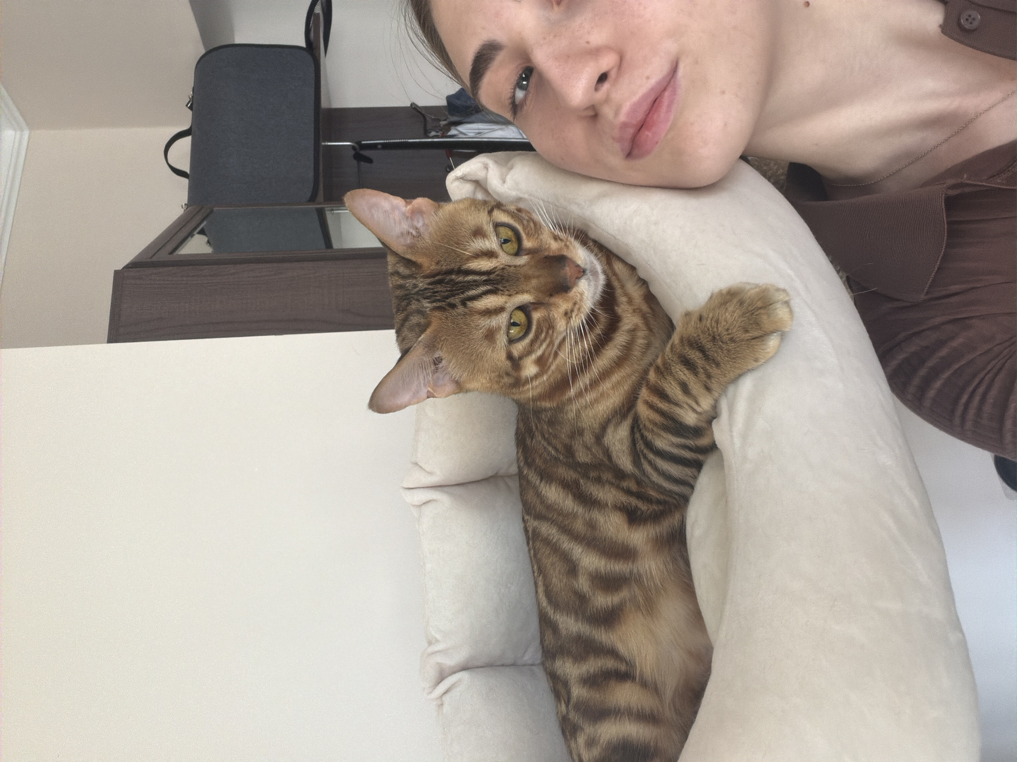 A young woman and a Bengal cat are in a selfie. The woman's face is partially visible on the right side of the frame, looking towards the camera. Her skin is fair with freckles, and she is wearing a brown collared shirt. A delicate silver necklace is visible. The cat, positioned on the left, is resting its head on a plush white cushion and is looking directly at the camera with intense yellow eyes. The cat has distinctive striped markings typical of its breed. The background suggests an indoor setting with a wardrobe and a dark grey bag on a shelf. The lighting is soft and natural. The location in Krasnoyarsk, Russia, cannot be determined from the image.
