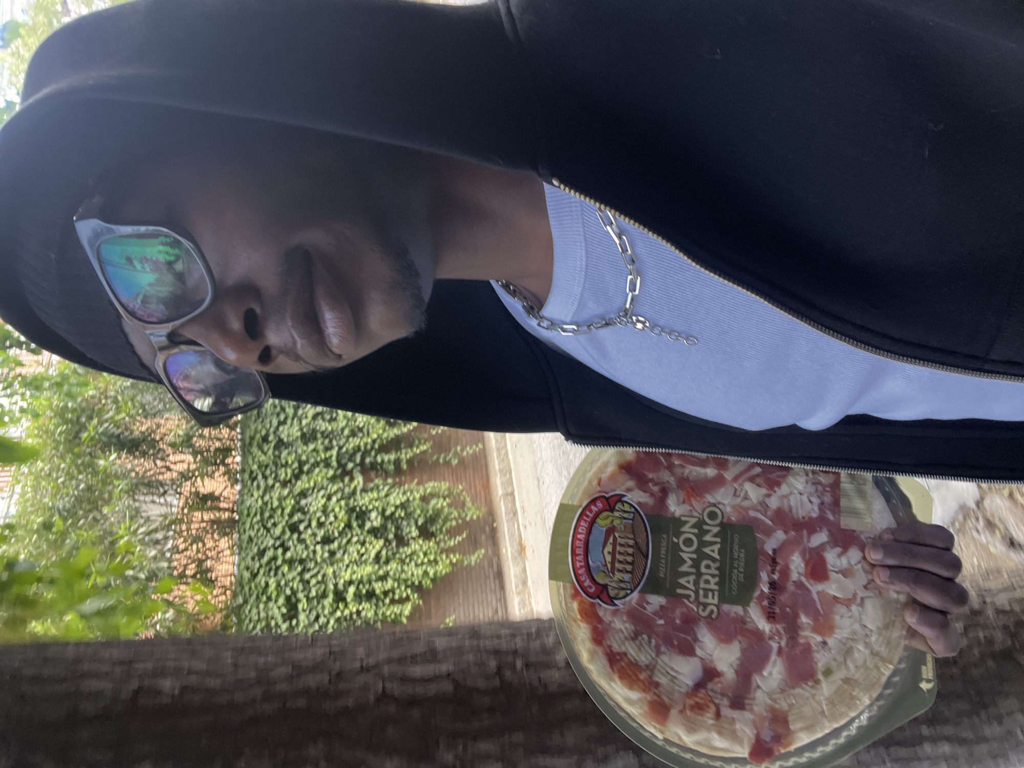 A person wearing a black hoodie, sunglasses, and a silver chain holds a packaged pizza with "Jamón Serrano" labeled on it. The person is outdoors with green vegetation and a brick wall in the background. It is likely a park in Barcelona due to the combination of Mediterranean vegetation and brick walls being a common sight in Barcelona.