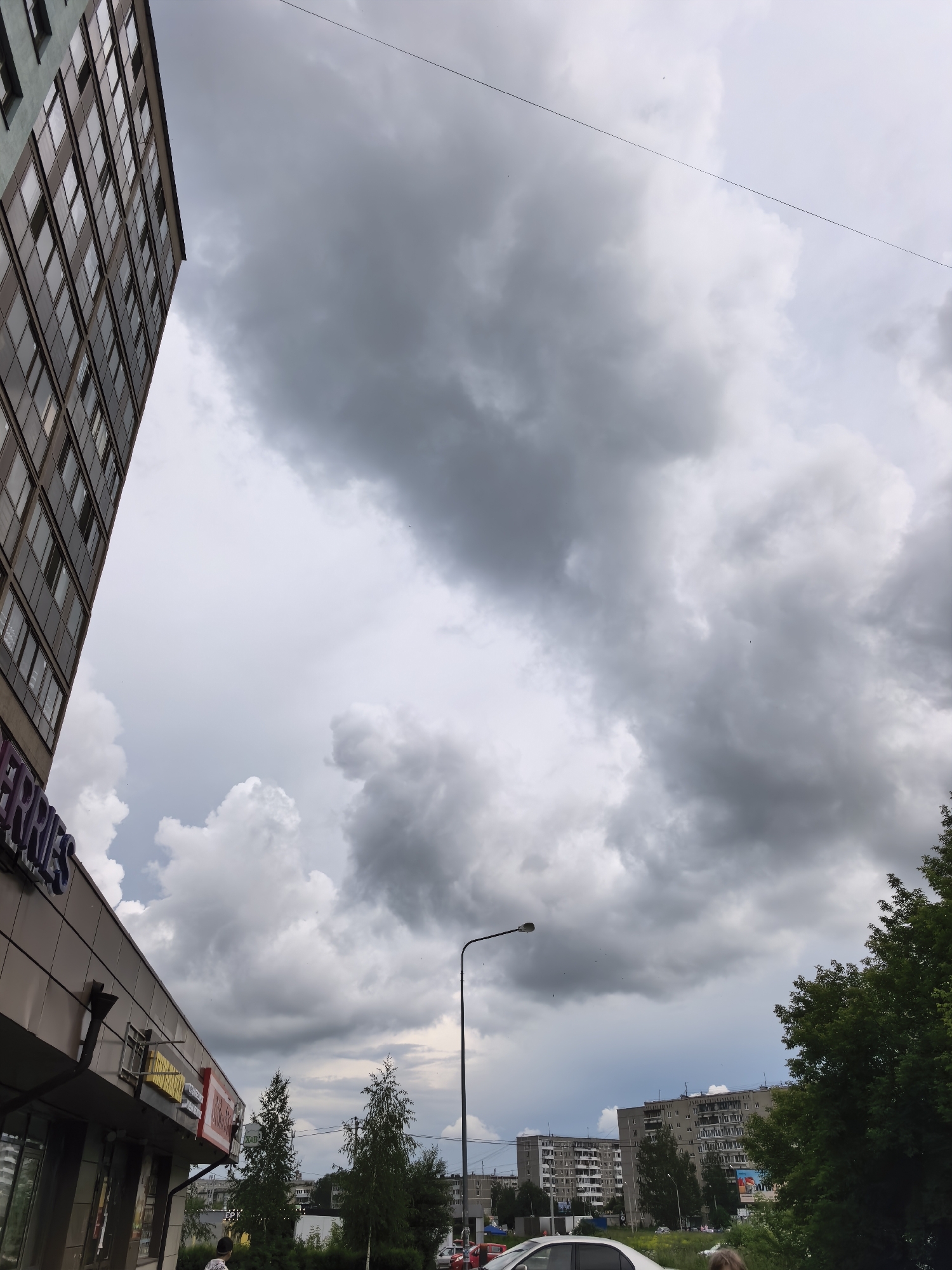 The image shows a street-level view in an urban environment, dominated by a dramatic, overcast sky filled with dark, heavy clouds. In the foreground on the left, the side of a modern, multi-story building is visible, featuring a facade with numerous windows and a dark blue sign reading "BERRIES". Below this, there are illuminated signs with yellow and red elements. To the right of the building, a street lamp stands tall. Further into the background, several tall, residential apartment buildings with many windows are visible, some appearing to be older Soviet-era structures. Green trees and bushes line the street and areas between buildings. A light-colored car is parked on the right side of the road, and other vehicles are visible in the distance. The overall scene suggests an impending change in weather, possibly rain, due to the dark clouds.