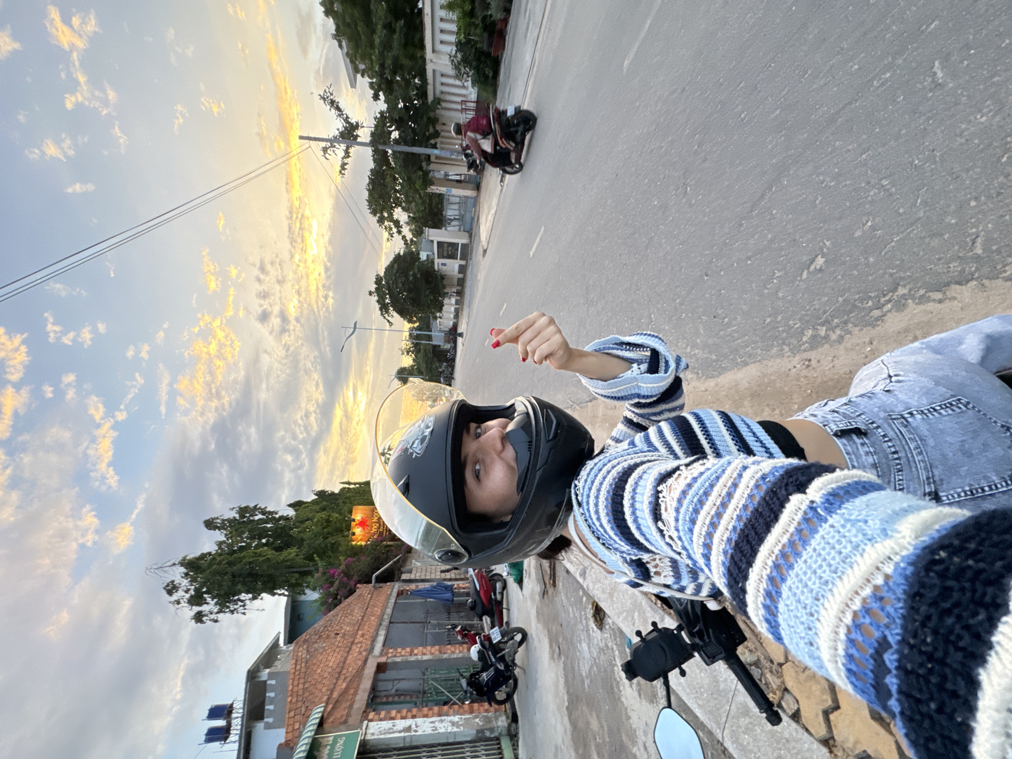 A woman wearing a black helmet and a blue and white striped sweater is sitting on a motorcycle. She is smiling and making a heart shape with her fingers. The motorcycle is parked on the side of a road in Mũi Né, Vietnam. In the background, there are buildings, trees, and a cloudy sky at sunset. Several other motorcycles are also visible on the road and parked along the street. The scene appears to be a street in Mũi Né, Vietnam, during the golden hour.