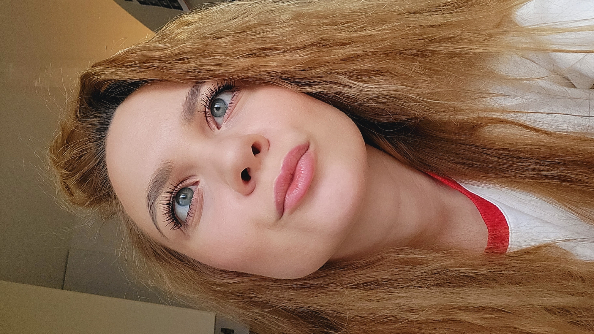A close-up, portrait-style image features a young woman with light skin, light blonde/auburn hair, and light blue eyes. She is facing the camera but is tilted slightly to the left. Her hair is long and wavy, framing her face. She is wearing makeup, including noticeable mascara on her eyelashes. A small portion of a white shirt with a red trim is visible along her shoulder. The background is out of focus. This image was taken in the city of Chaikovsky, Russia.
