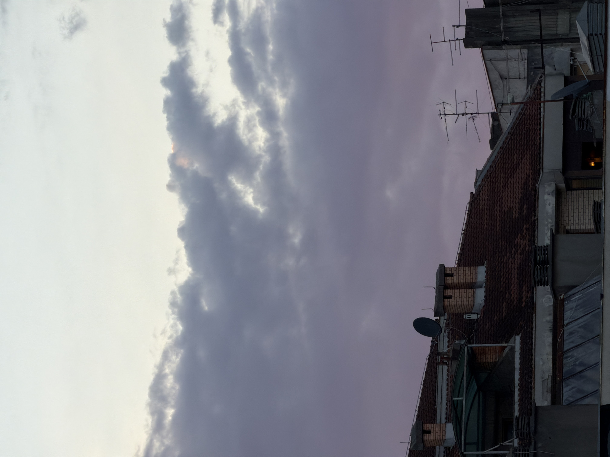 The image shows a view of rooftops and the sky at dusk in Podgorica, Montenegro. The sky is filled with heavy, dark clouds that have a purple hue, indicating twilight. There is a sliver of lighter sky visible at the top, suggesting the sun may have recently set or is about to rise.
On the rooftops, several architectural elements are visible. These include:
* **Roofs:** Predominantly tiled with reddish-brown tiles.
* **Chimneys:** Several brick chimneys of varying sizes and designs are present. Some have multiple openings.
* **Satellite Dishes and Antennas:** A dark, parabolic satellite dish is mounted on one of the roofs. Additionally, several television antennas, including a large, multi-pronged one, are visible.
* **Structural Elements:** Other parts of buildings, including concrete structures and glass-covered sections, are also visible.
* **Lighting:** A faint yellow light emanates from a window in one of the buildings, suggesting an interior light.
The scene is devoid of any visible people or animals. The overall impression is that of an urban landscape at the end of the day, with a somewhat dramatic sky. The composition focuses on the textures and shapes of the rooftops and the atmospheric conditions of the sky.