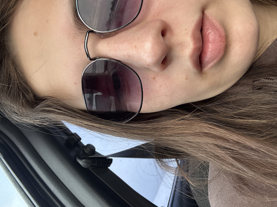 The image is a close-up portrait of a person, captured from the forehead down to the chin, with the subject's head significantly tilted to the left. The person has fair skin with visible freckles scattered across their nose and cheeks. They are wearing sunglasses with dark, possibly reflective lenses and a thin metal frame, covering their eyes and upper face. The bridge of the sunglasses rests on their nose. The person's lips are a natural light pink or peach color and are slightly parted. Long, light brown or auburn hair, appearing smooth, frames their face and extends towards the bottom and right edges of the frame.
In the lower left corner of the image, parts of a car interior are visible, featuring a black textured surface and a black plastic clip or mount. Beyond this car element, a blurry, light-colored area suggests an outdoor background, possibly the sky. A small section of a light brown or beige garment, likely a shirt or jacket, is visible on the right side of the image. Due to the sunglasses, no specific emotions are discernible. The presence of sunglasses indicates it is likely daytime. No text is visible in the image.